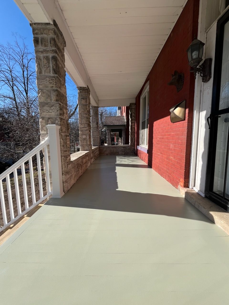 A covered porch with a light-colored floor, brick wall, stone columns, and white railing.