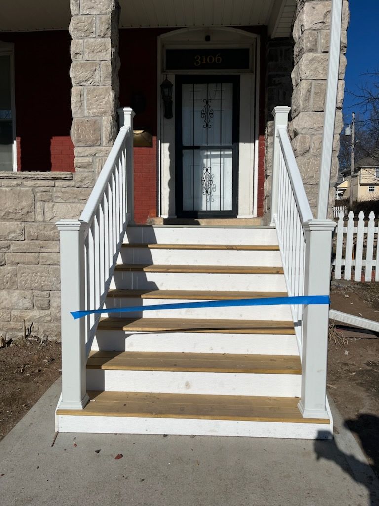 A front porch with white railings and wooden stairs leading to a black door, blue tape across the steps.
