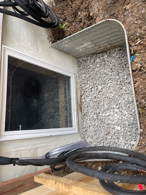 Window well with gravel, next to a basement window. Black cable in the foreground and soil in background.