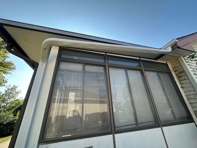 Low-angle view of a screened porch with dark-framed windows and white trim, under a roof with a gutter.