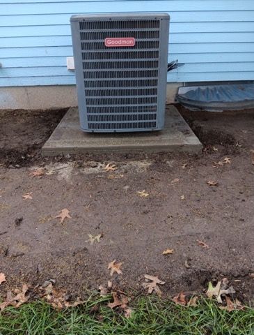 Air conditioner unit on concrete pad next to a blue house, surrounded by dirt and leaves.