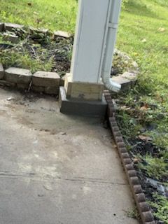 Concrete porch post with downspout, bordered by bricks and lawn.