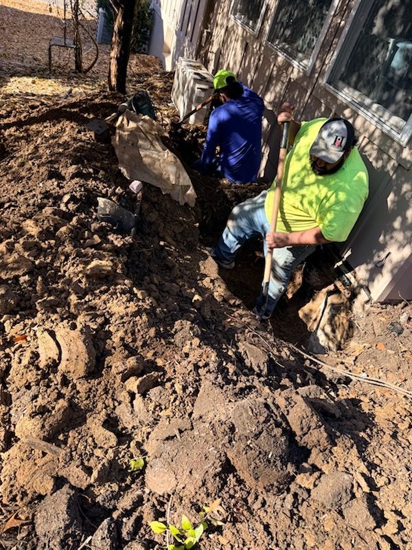 Two workers digging a trench next to a building. One wears a green shirt, the other blue. Soil visible.