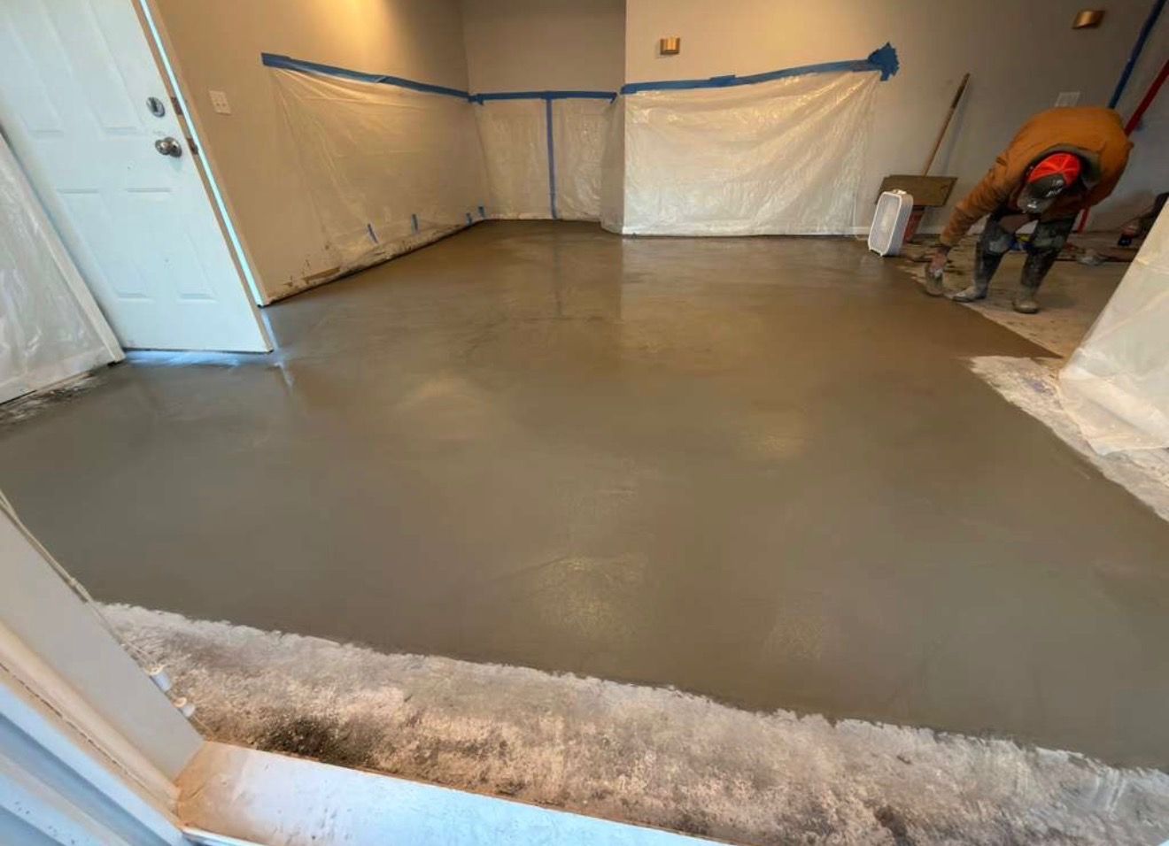 A worker smoothing wet concrete flooring in a room, walls covered in protective plastic.