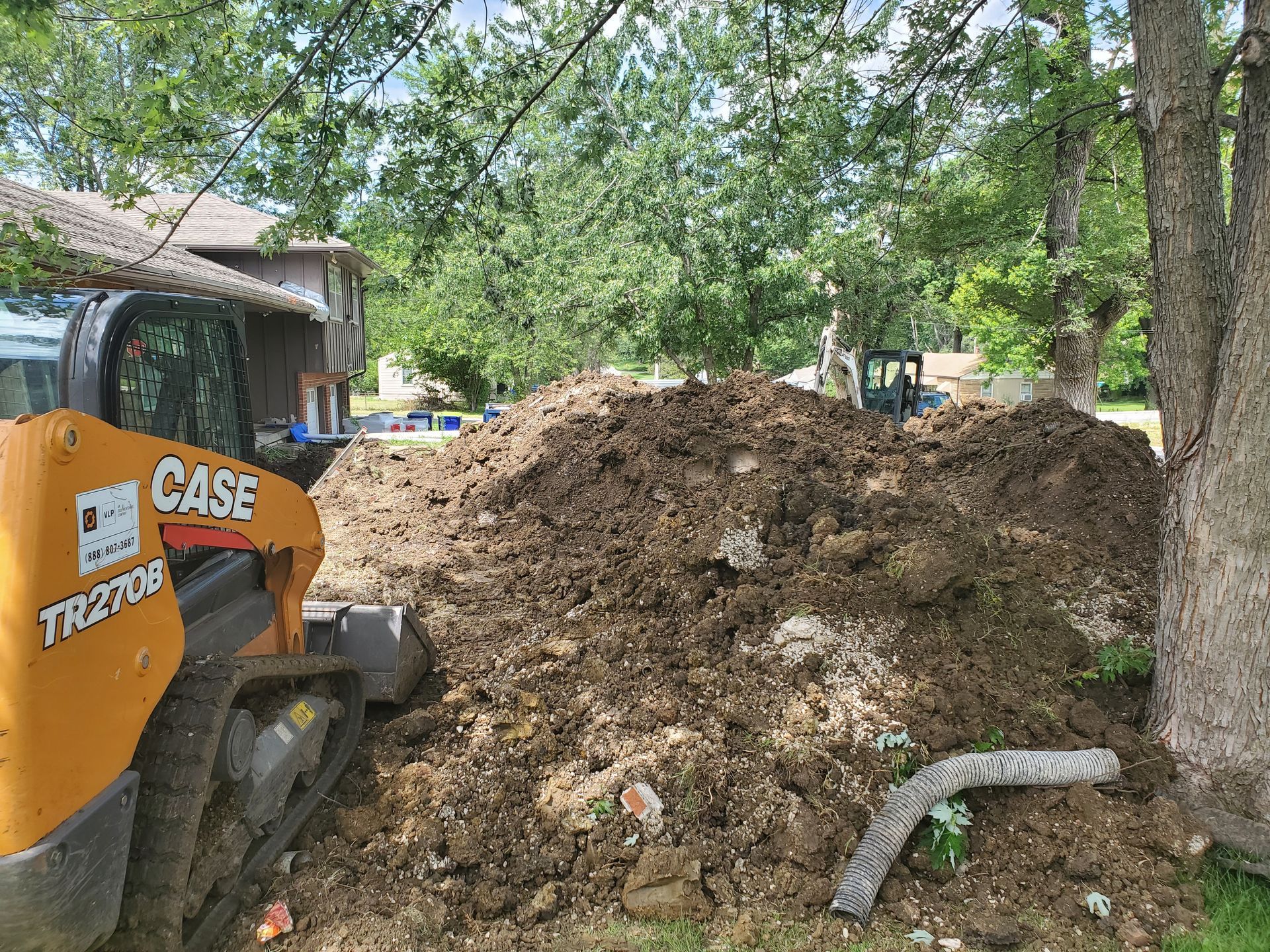 A Case TR270B skid steer next to a large pile of dirt, near a house and trees.