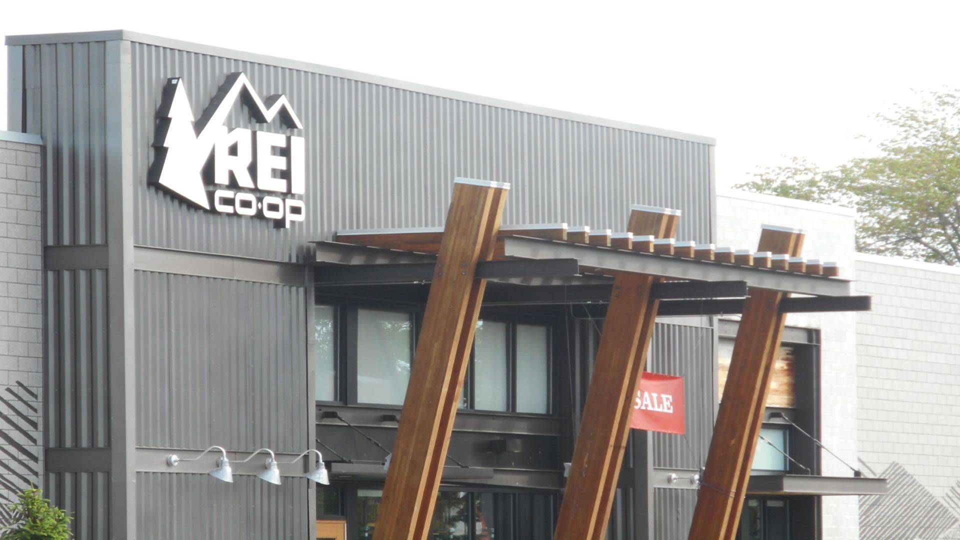 REI Co-op store exterior with logo, gray corrugated metal siding, wooden supports, and a red sale banner.