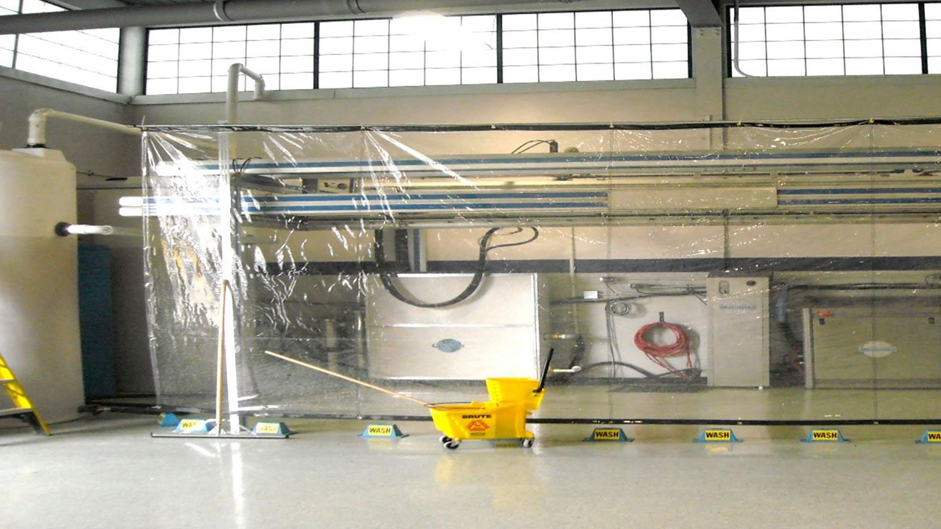 Inside a factory, a clear plastic barrier surrounds equipment; a yellow mop bucket is present on the floor.
