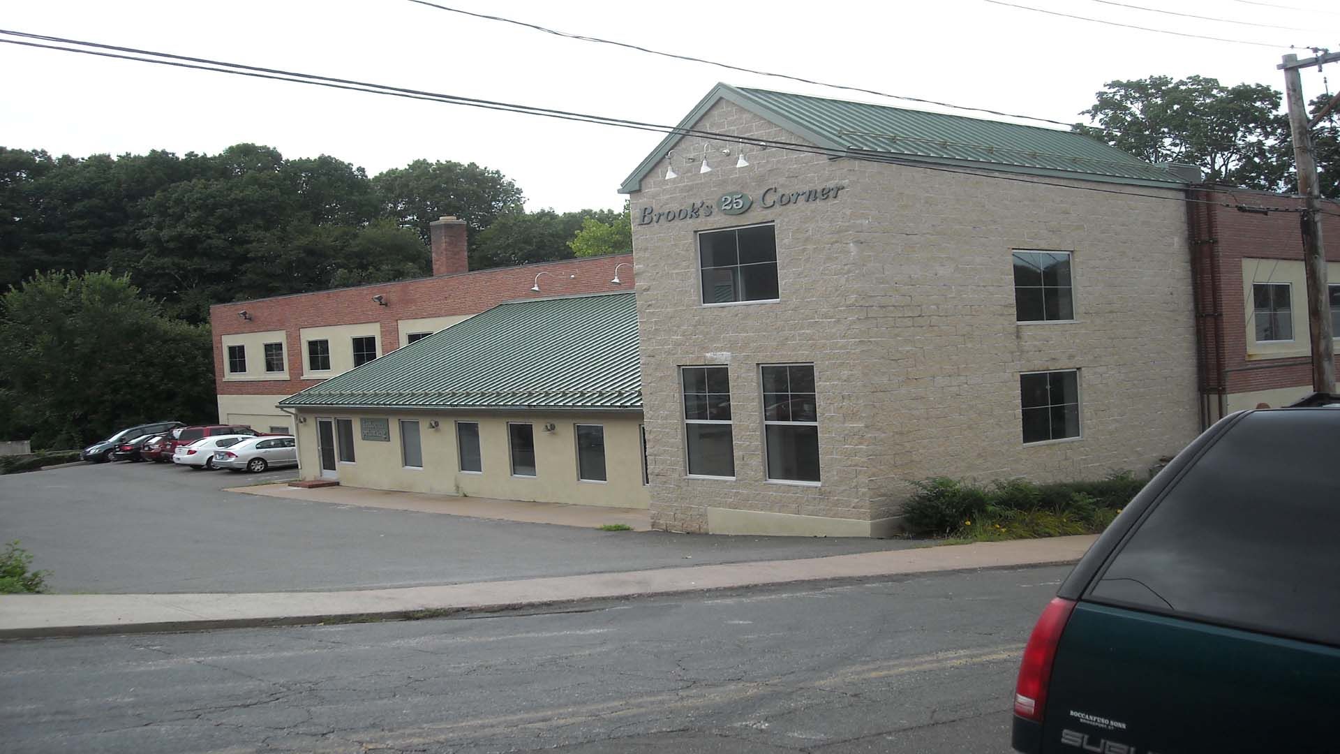 A light-colored brick building with a green tiled roof, parked vehicles, and a gray sky.