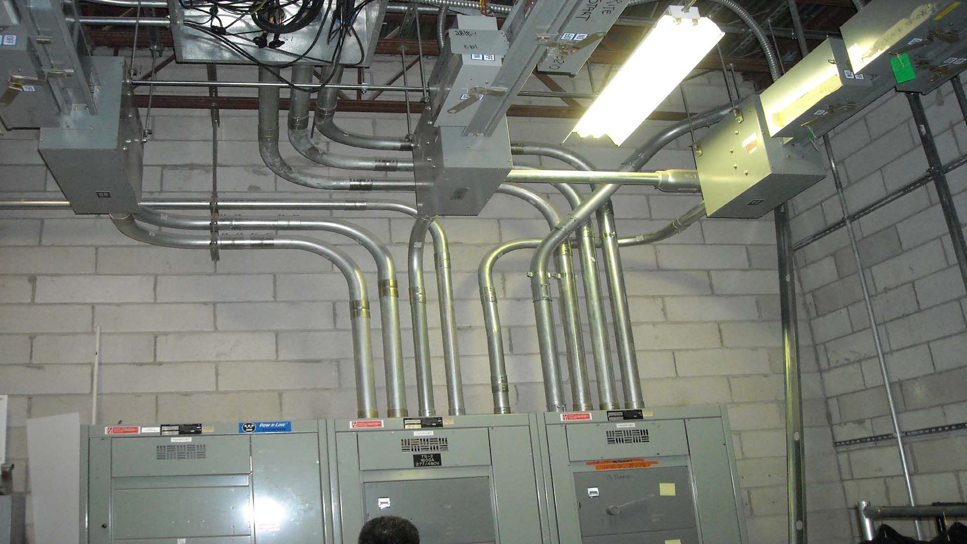Electrical conduit and junction boxes run along a brick wall above electrical panels.