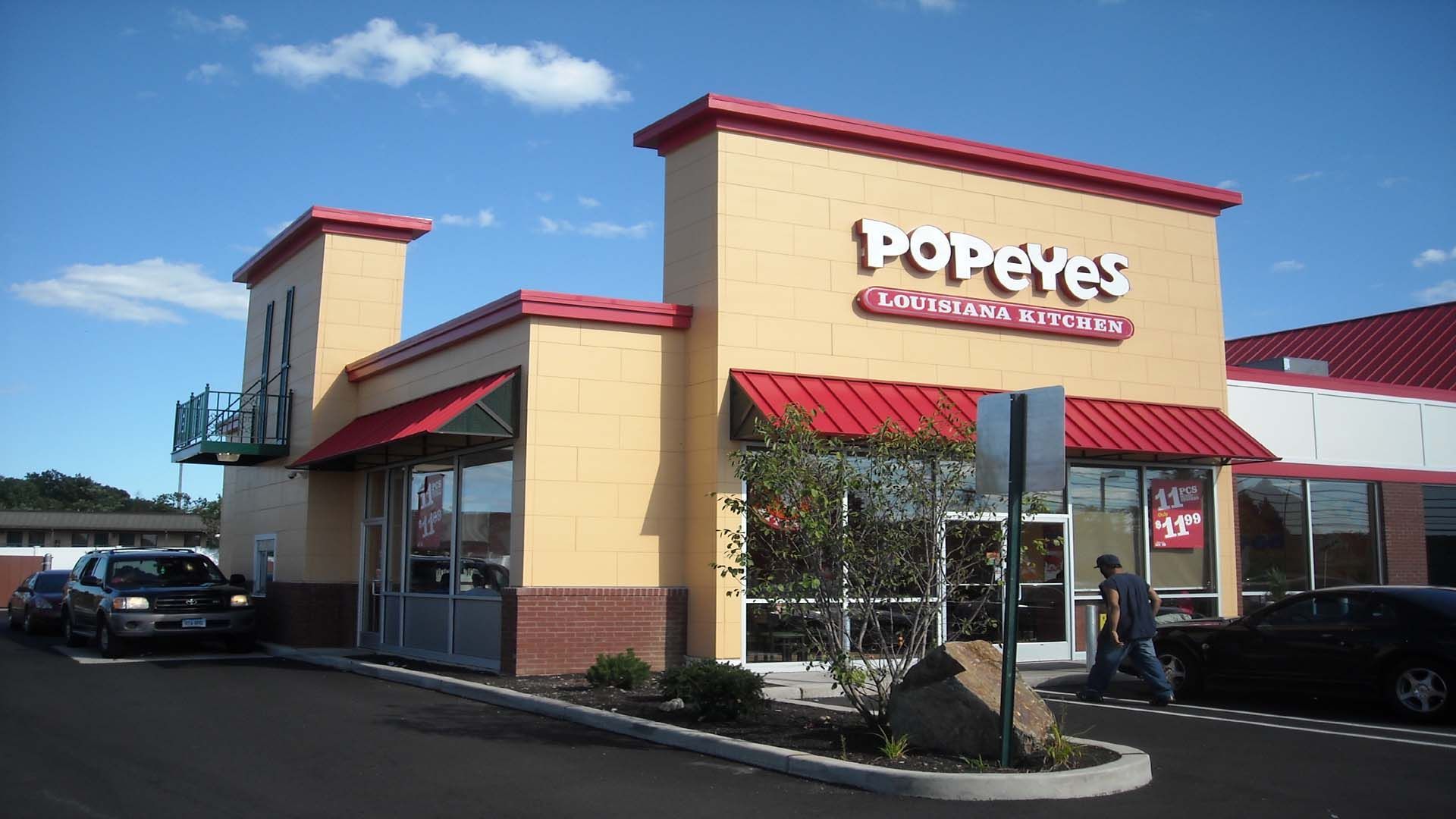 Popeyes restaurant exterior, beige with red accents, drive-thru, person entering, cars present.