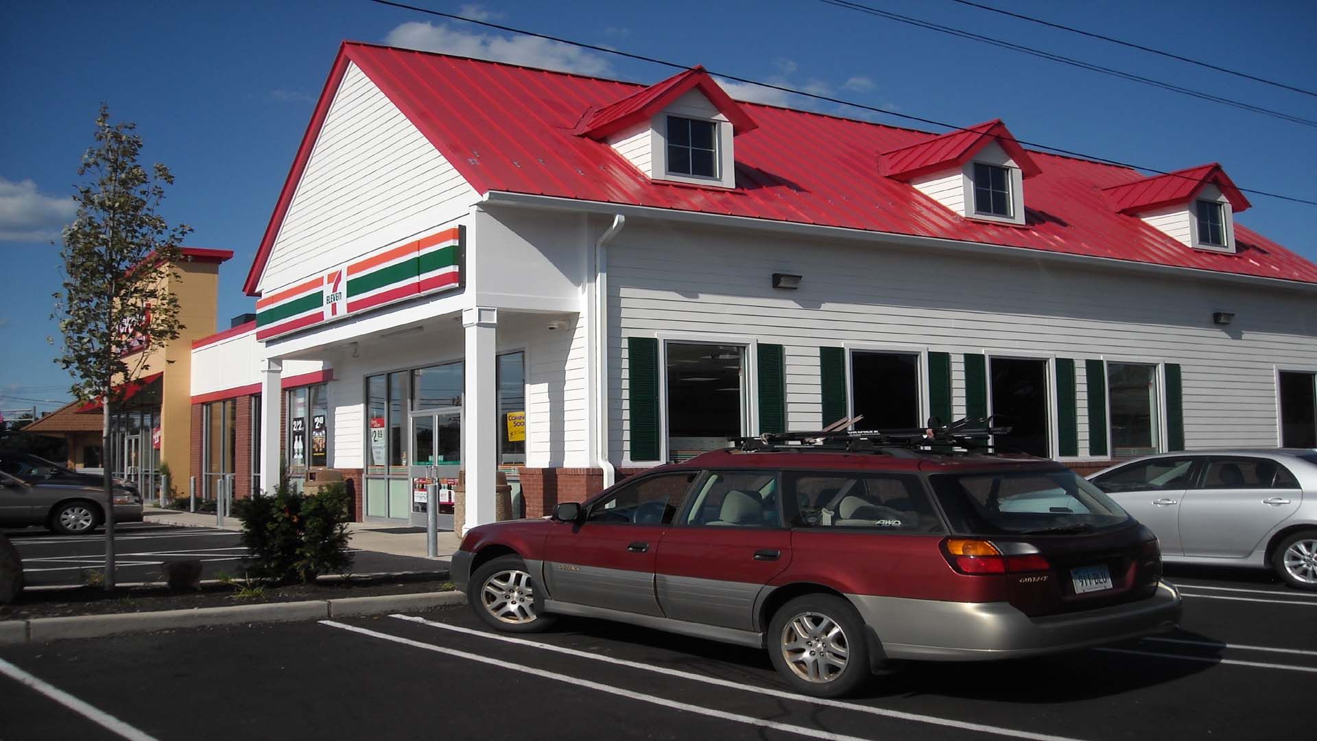 7-Eleven store with red roof, parked cars, and blue sky.