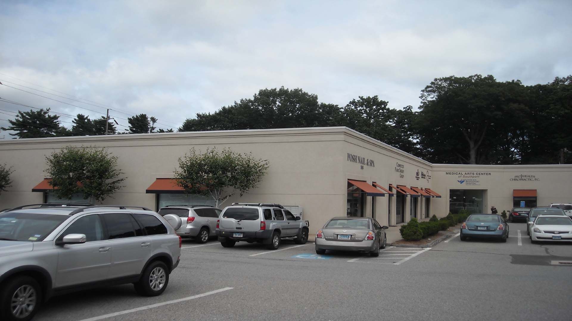 Cars parked in front of a cream-colored, single-story building with small orange awnings. Overcast sky.