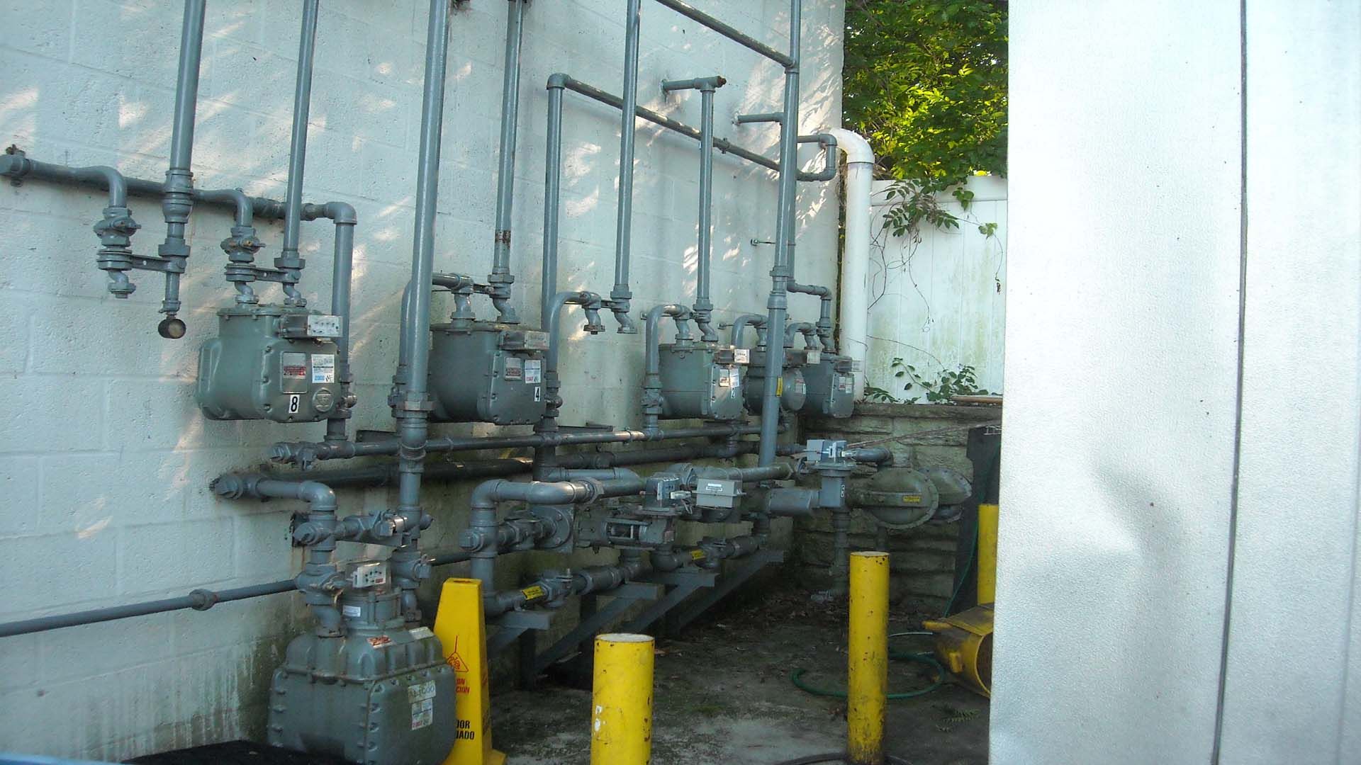 Gas meters and piping attached to a white building exterior, near yellow bollards.