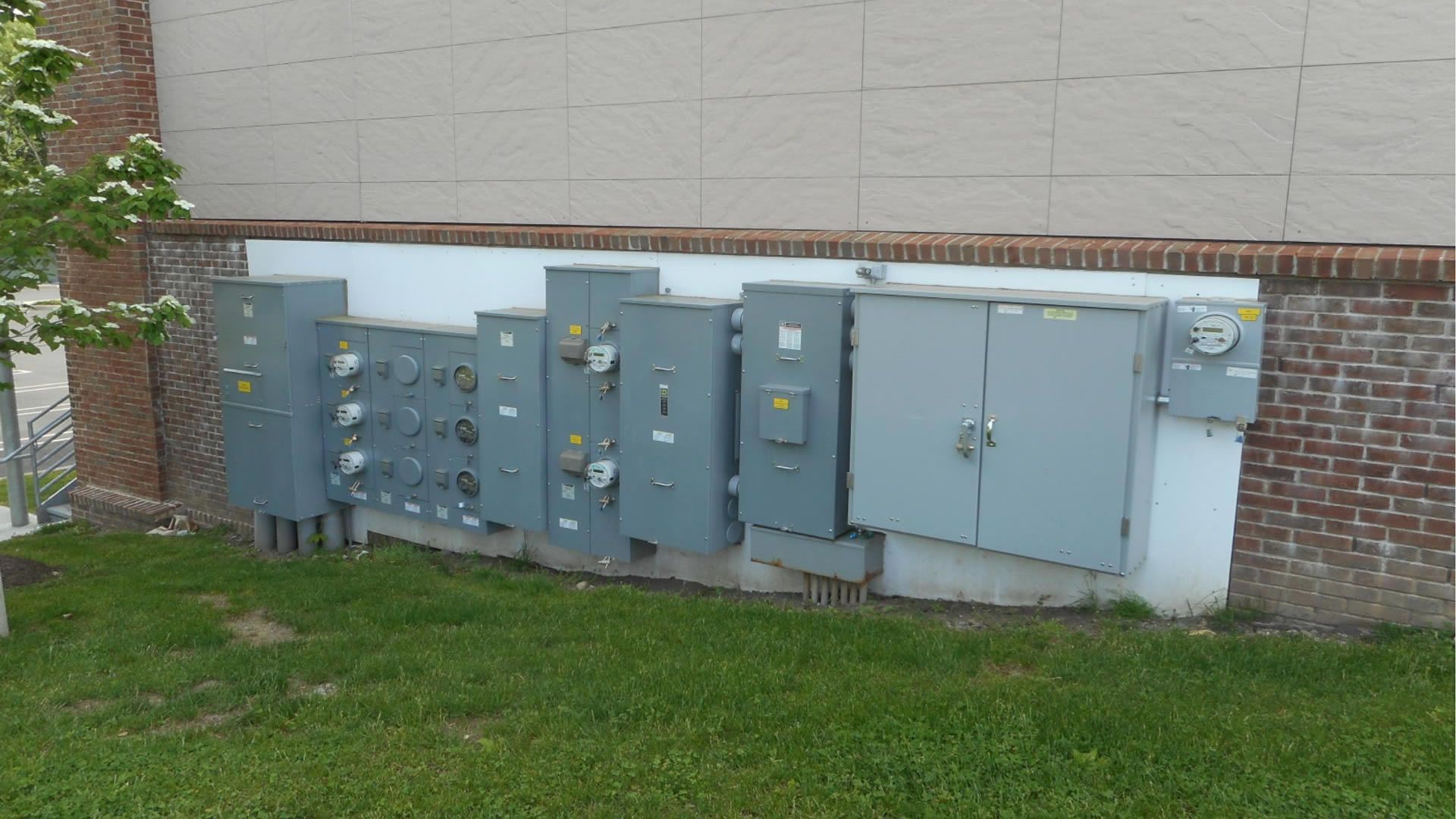 Electrical boxes mounted on a brick wall, next to a grassy area and a light-colored building.