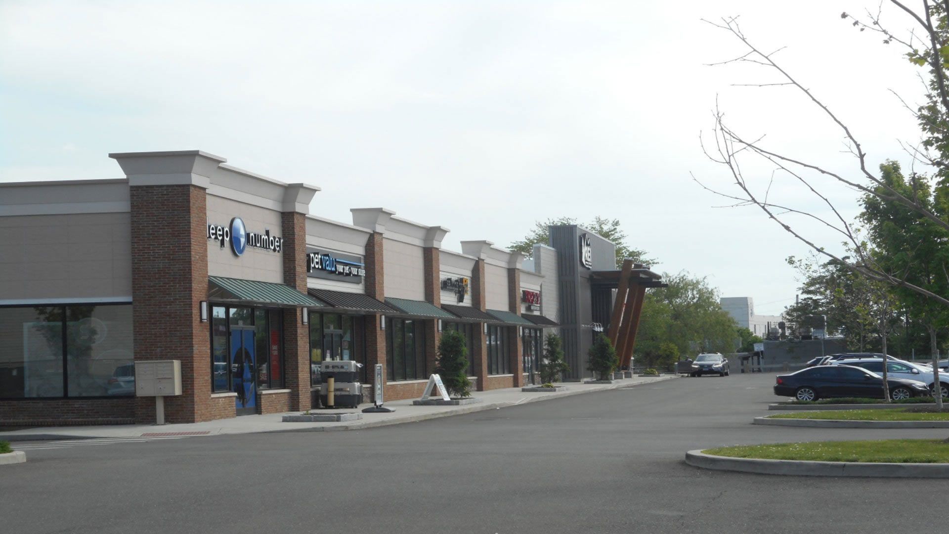 Strip mall with tan and brick facades, awnings, a road, and parked cars.