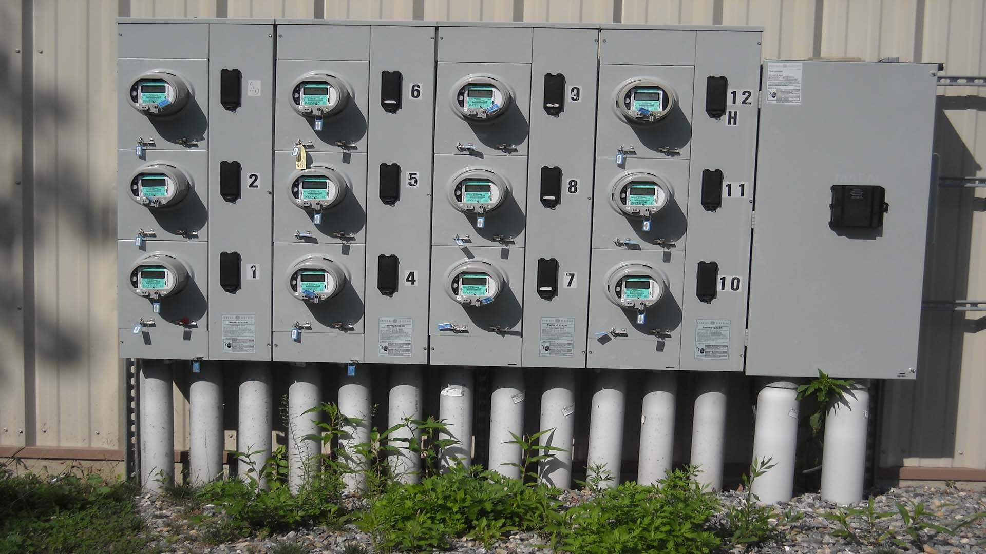 Gray electrical meter bank with multiple meters, numbered, mounted on concrete pillars against a wall.