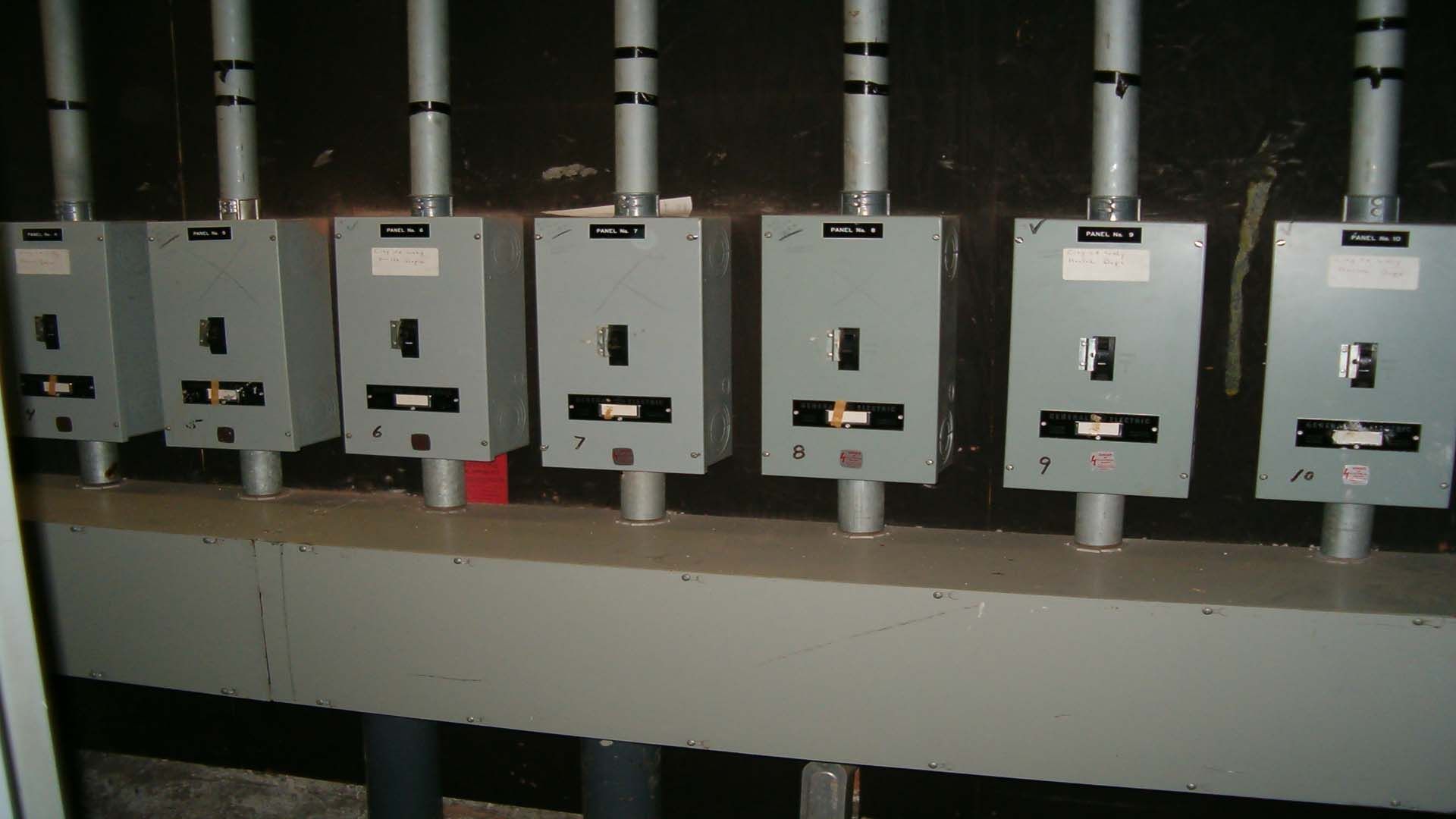 Seven gray electrical boxes with labels and conduits, mounted on a gray ledge against a dark wall.