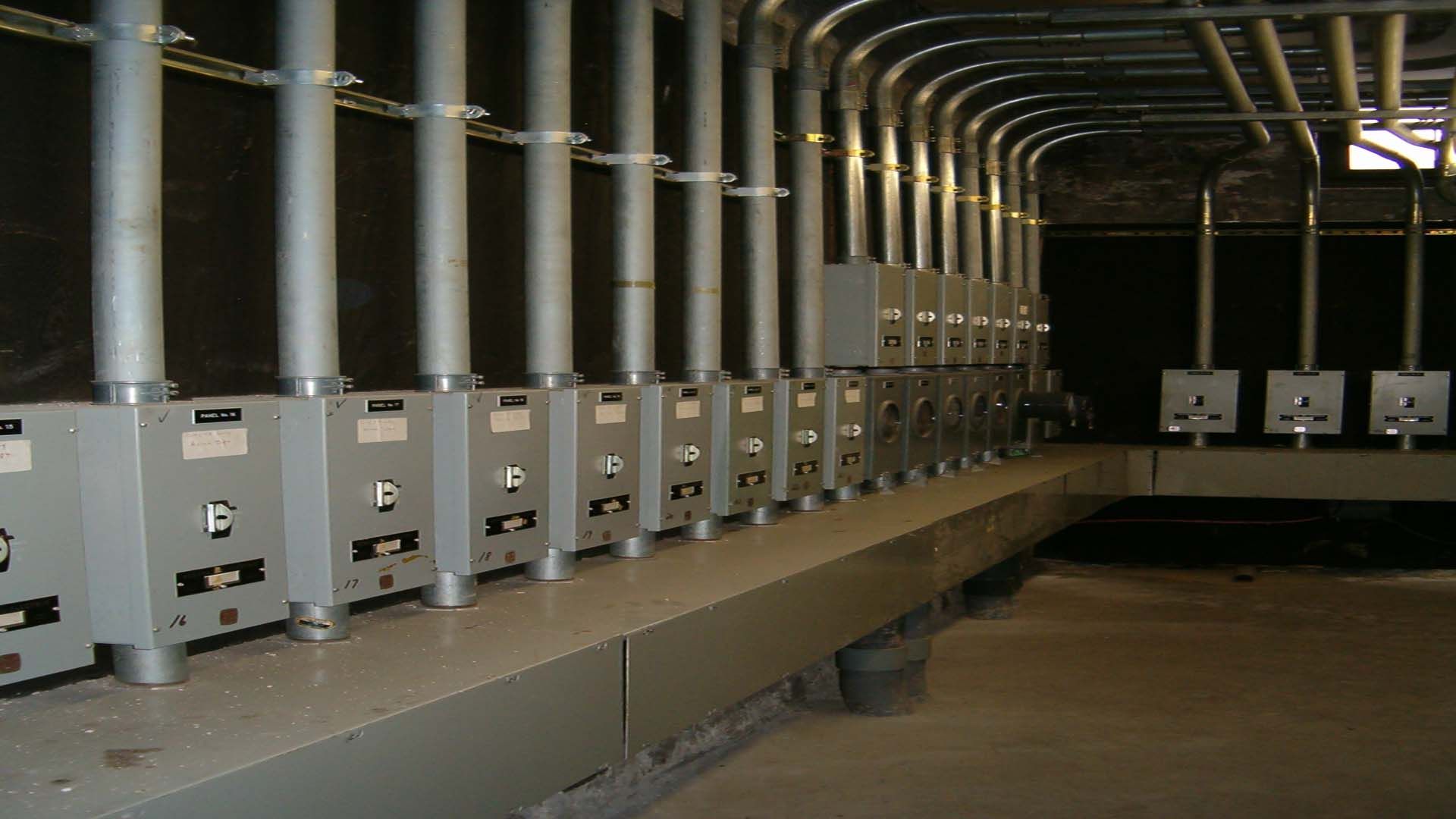 Gray electrical boxes and conduit lined along a wall, in an industrial setting.