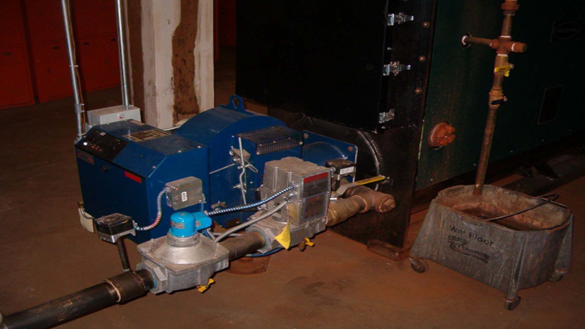Blue and silver furnace connected to a black industrial heating system; the furnace is in a basement.