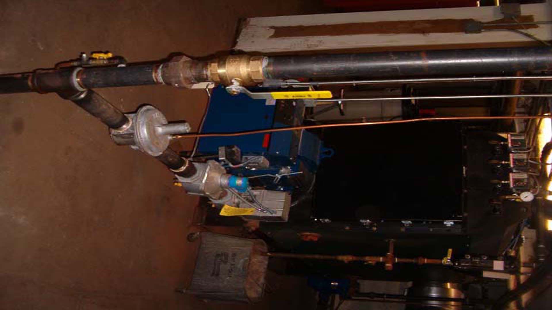 A large industrial boiler with black pipes, valves, and regulators in a utility room.