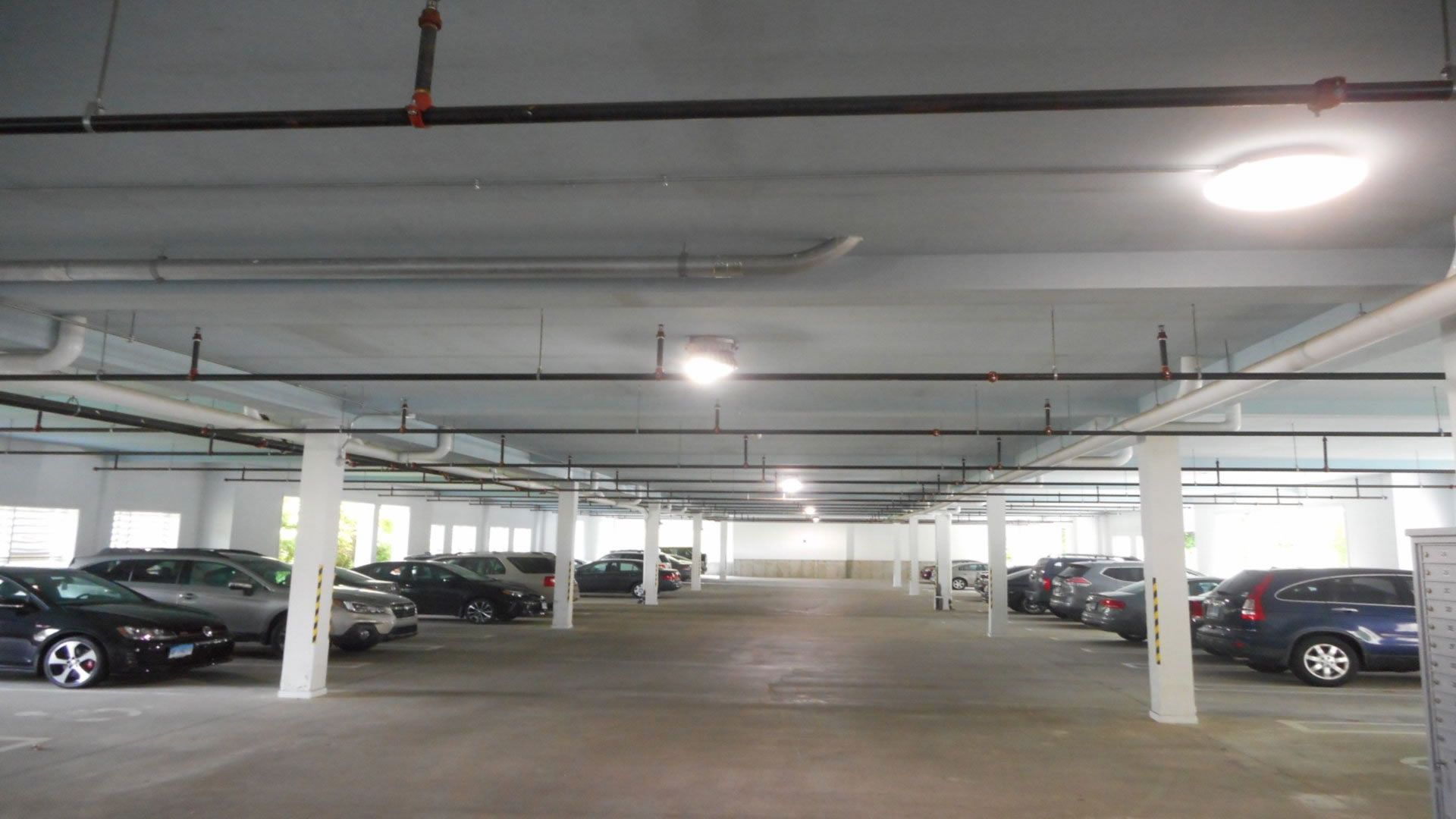 Indoor parking garage with rows of cars parked on both sides, overhead pipes and lights.