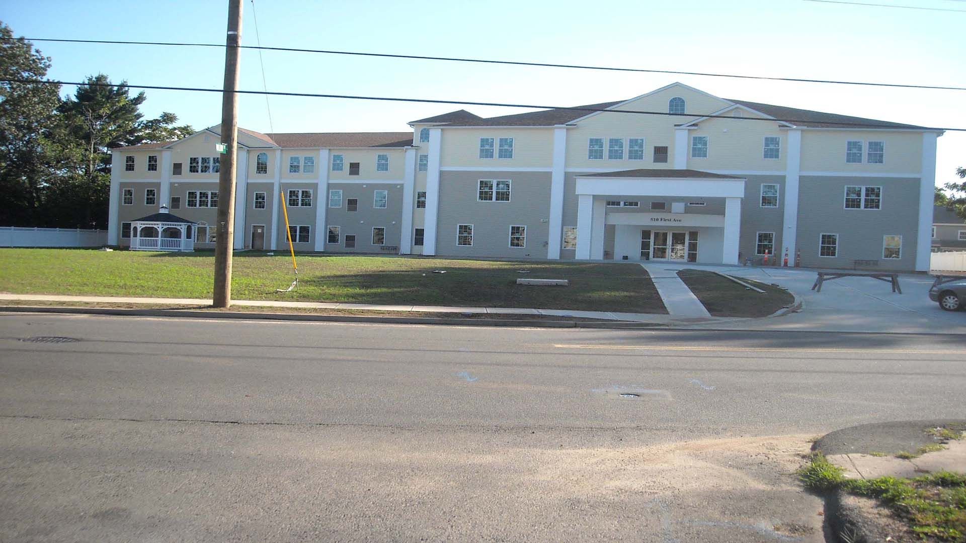 A multi-story light blue and gray building with a grassy lawn and a small gazebo; a street in front.