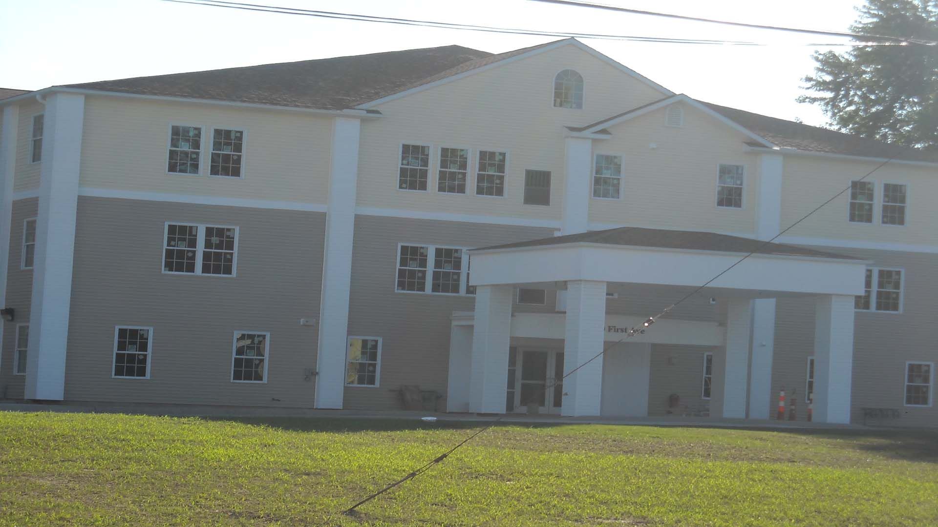 Three-story building with white pillars at the entrance, light yellow and grey exterior, grassy lawn.