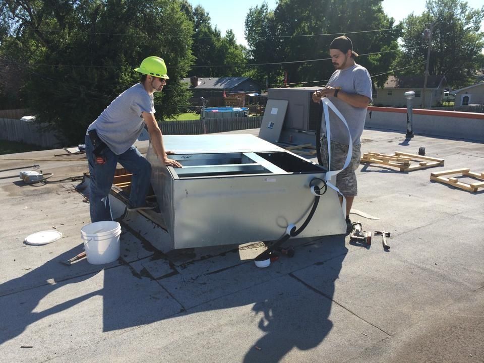 Two workers in gray shirts install a metal HVAC unit on a flat roof on a sunny day.