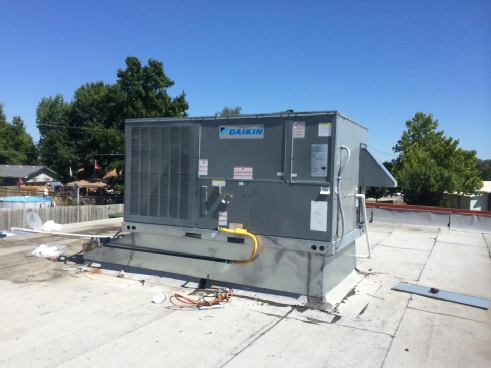 A gray Daikin HVAC rooftop unit sits on a flat commercial roof under a clear blue sky with trees in the background.