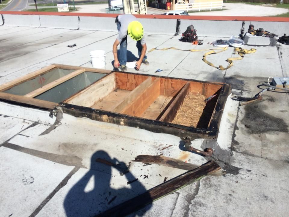 A worker in a yellow hard hat repairs a rectangular roof opening framed by wooden boards on a flat gray roof.