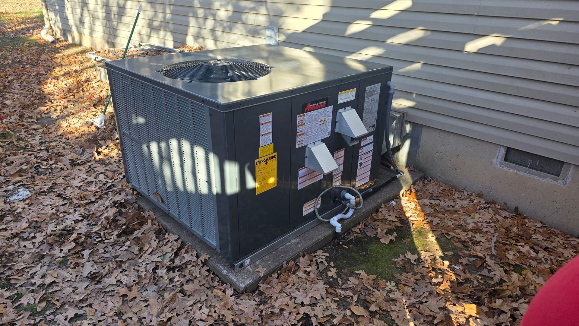A grey outdoor air conditioning unit sits on a concrete pad next to a house with beige siding, surrounded by dead leaves.
