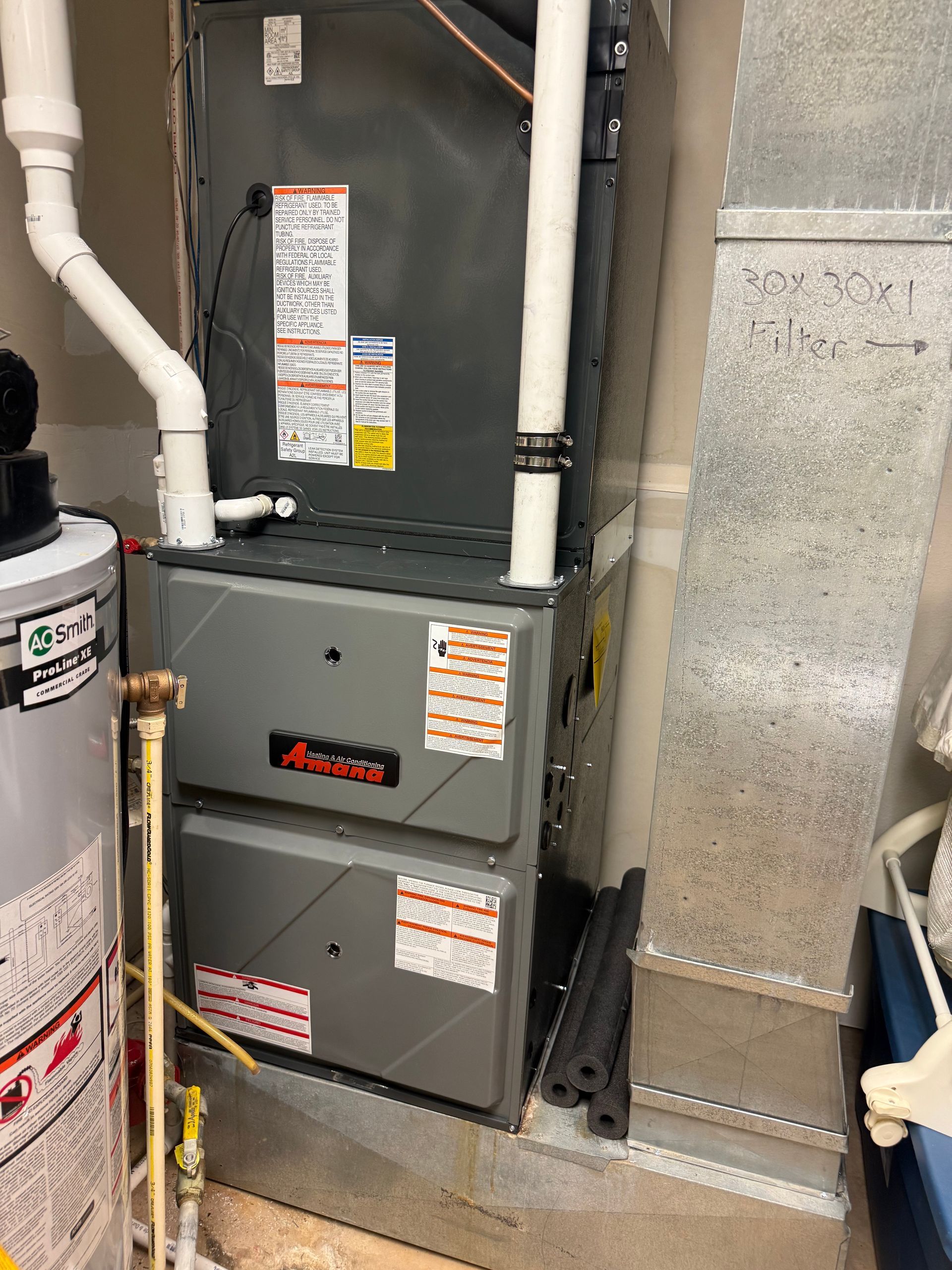 A dark grey HVAC furnace unit sits indoors next to a metallic duct, white PVC pipes, and a water heater.