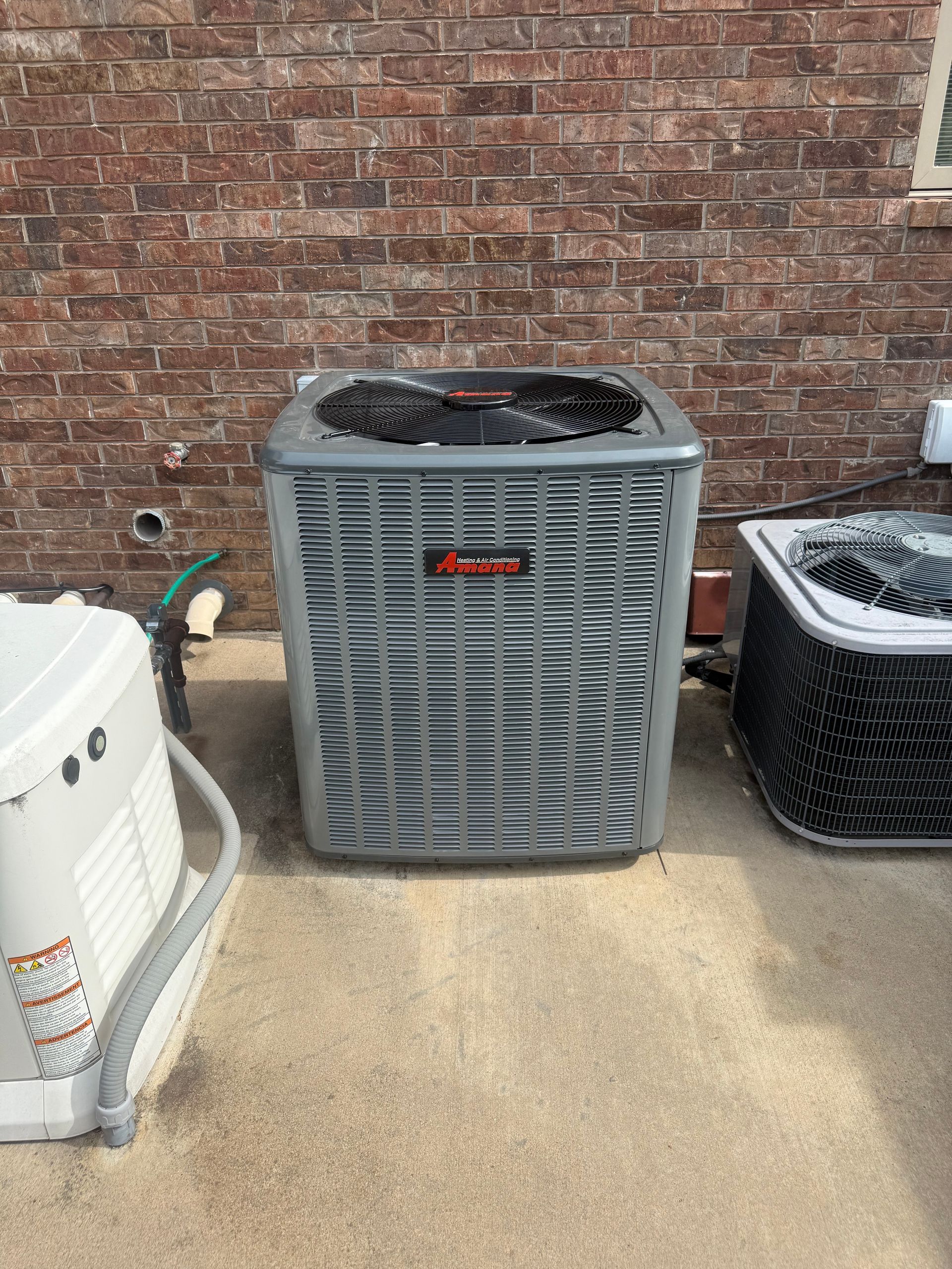 Three central air conditioning units sit on a concrete patio against a brick house exterior.