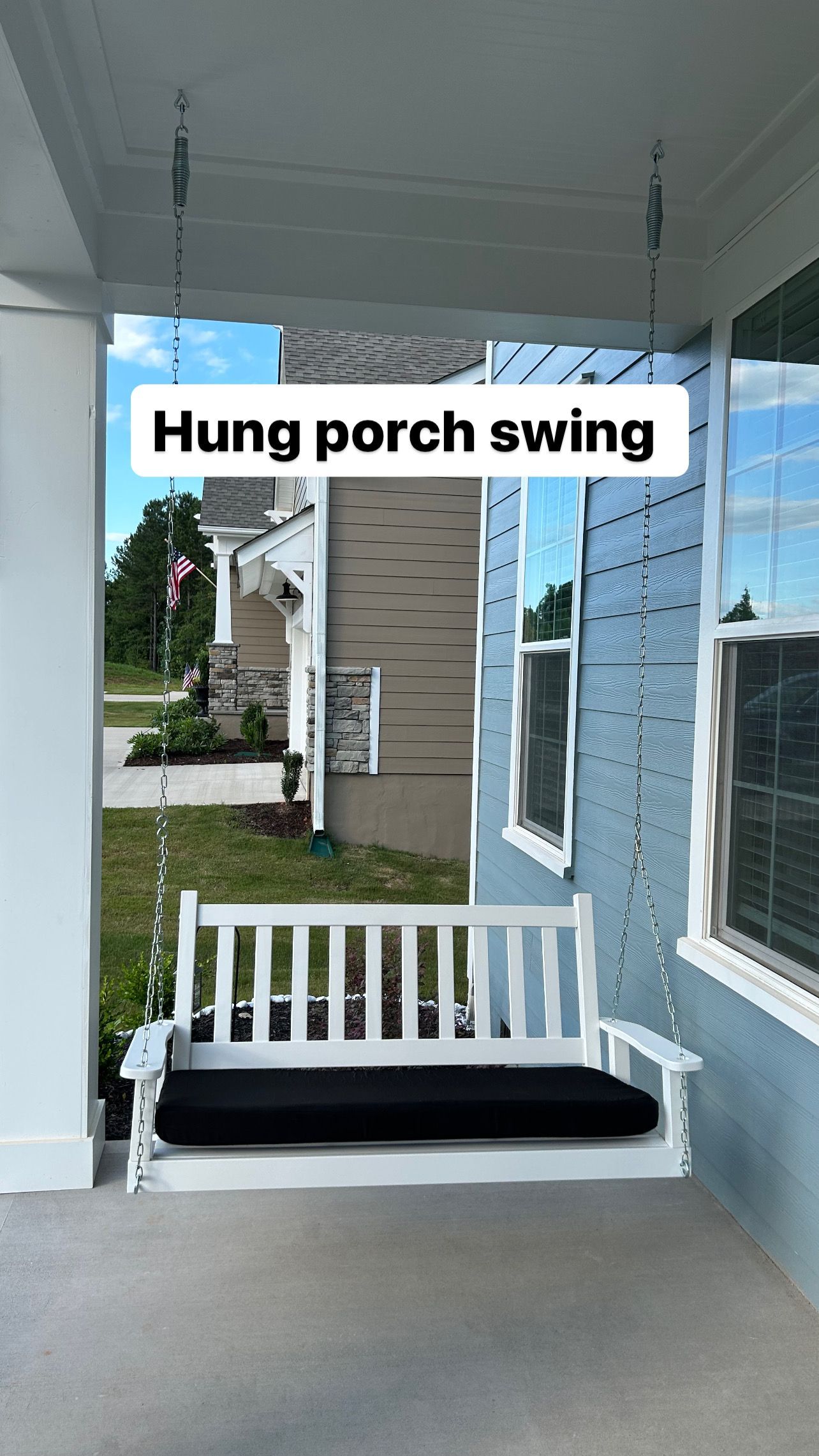 A white porch swing with a black cushion is hanging from the ceiling of a house.