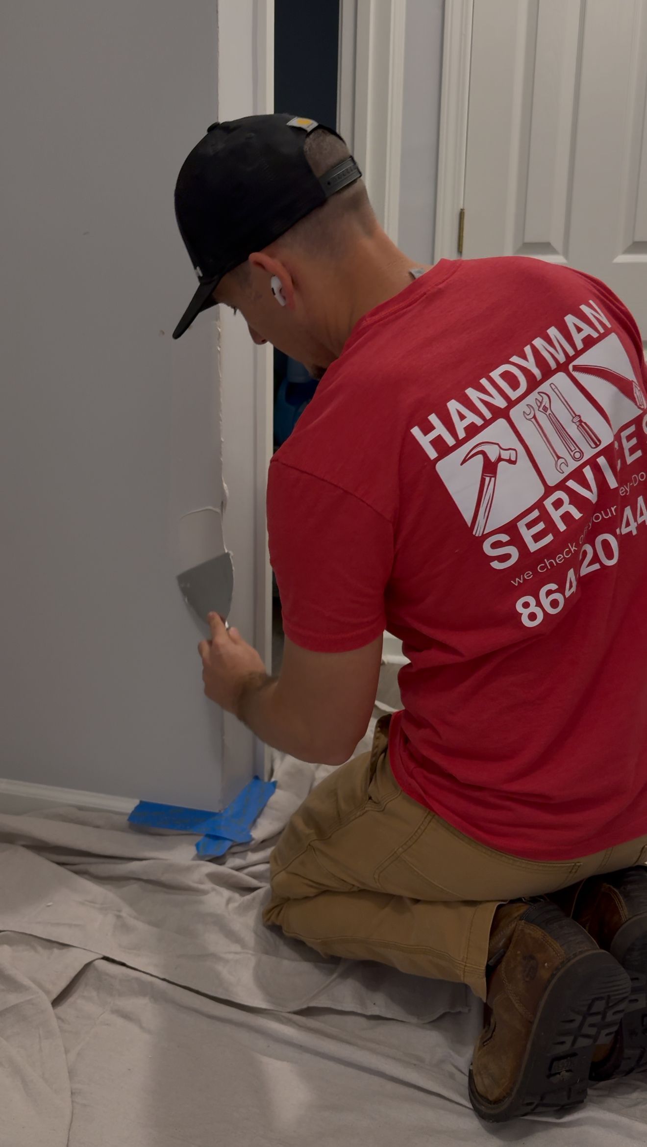 A man wearing a red handyman shirt is kneeling down and using a spatula on a wall.