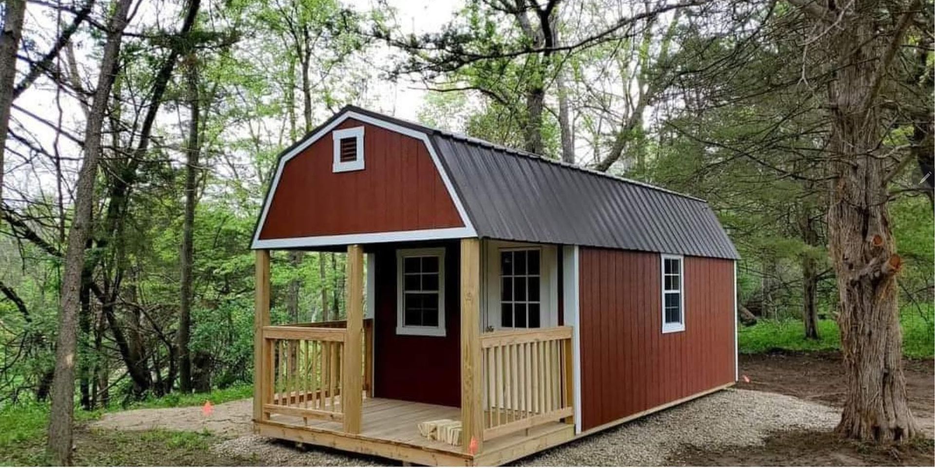 Small red cabin with a porch in a wooded area, with a gray metal roof and white-trimmed windows.