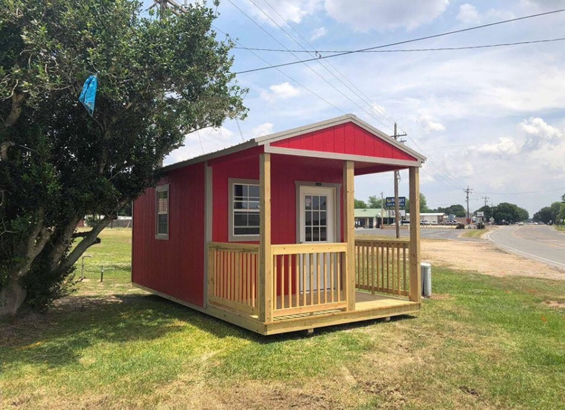 Small red cabin with a porch in a grassy roadside area under a partly cloudy sky