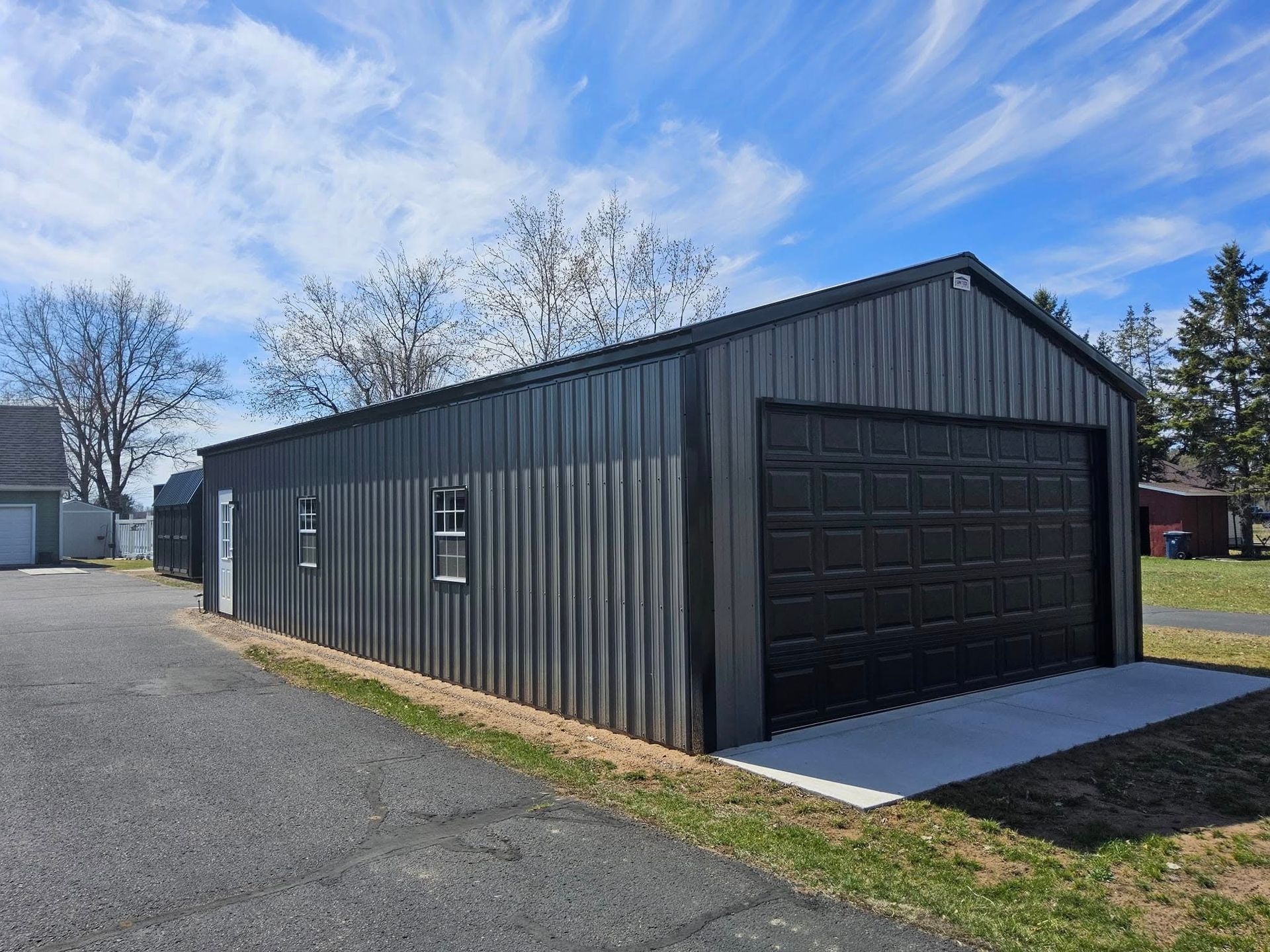 Dark gray metal garage with a black roll-up door beside a paved driveway under a blue sky