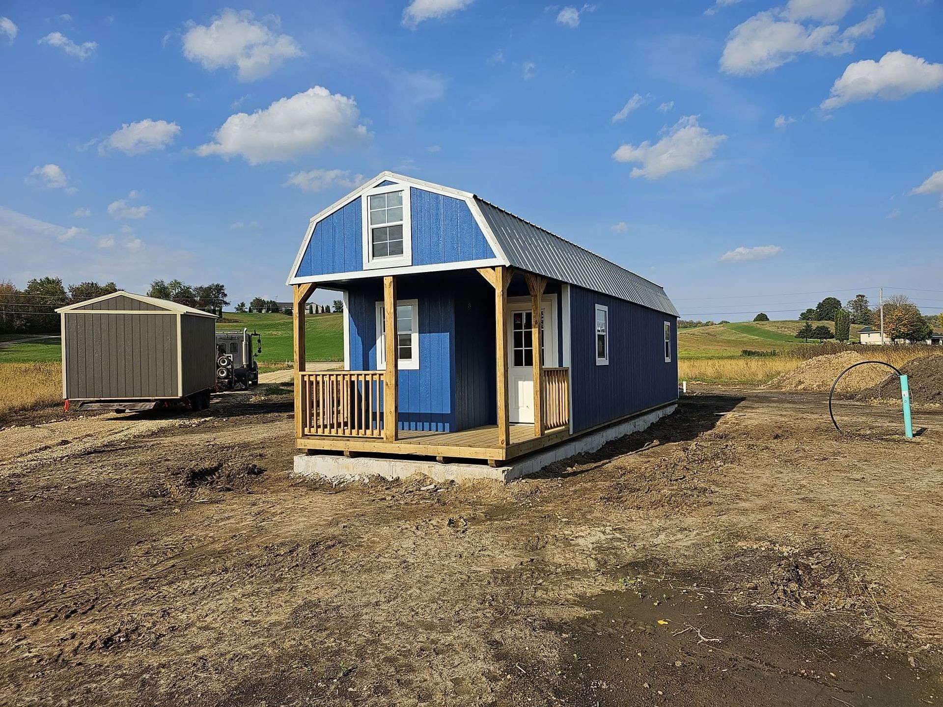 Small blue tiny house under construction on a dirt lot under a partly cloudy sky