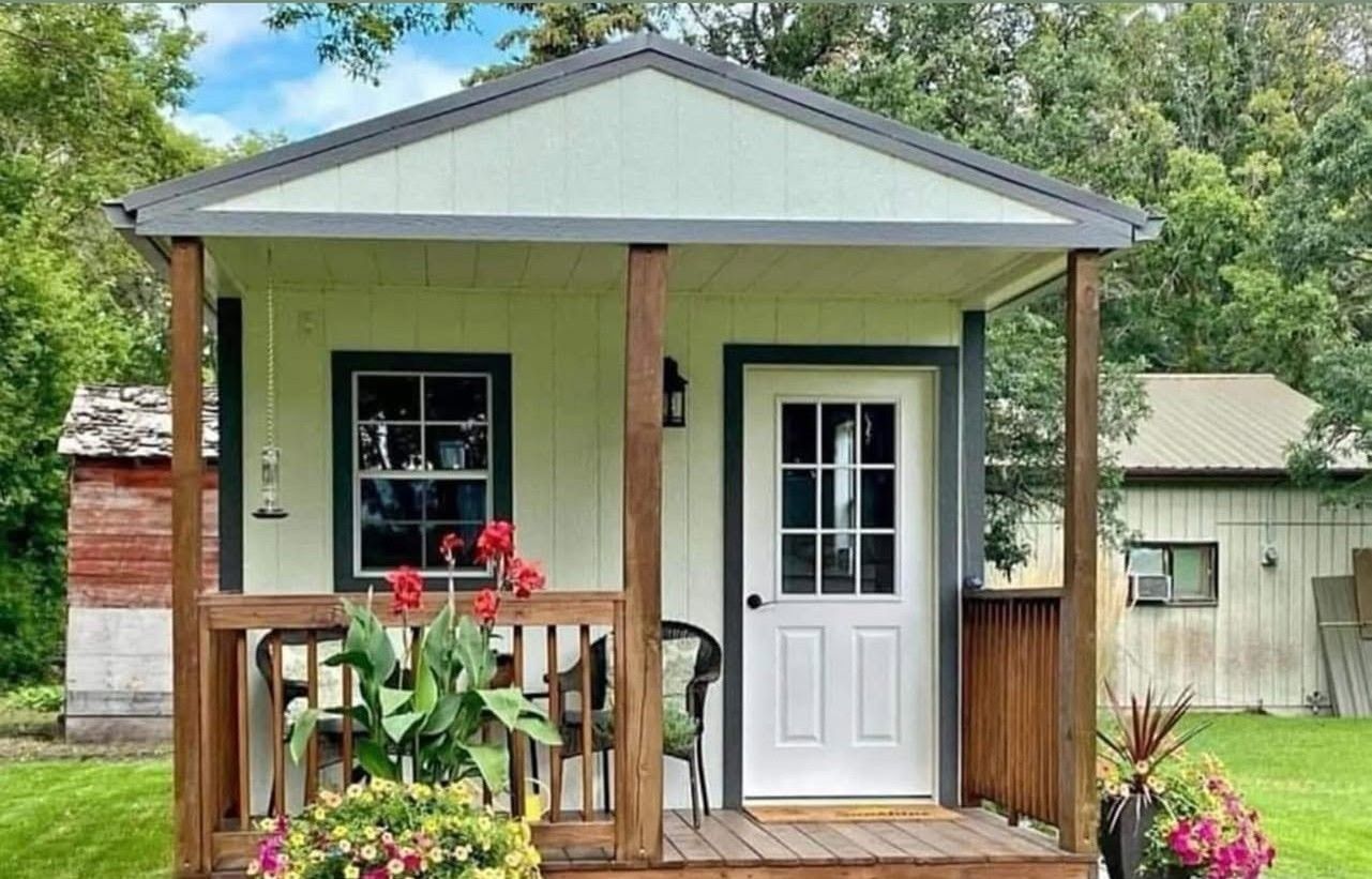 Small green front porch with white door, potted flowers, and a wooden railing in a yard.