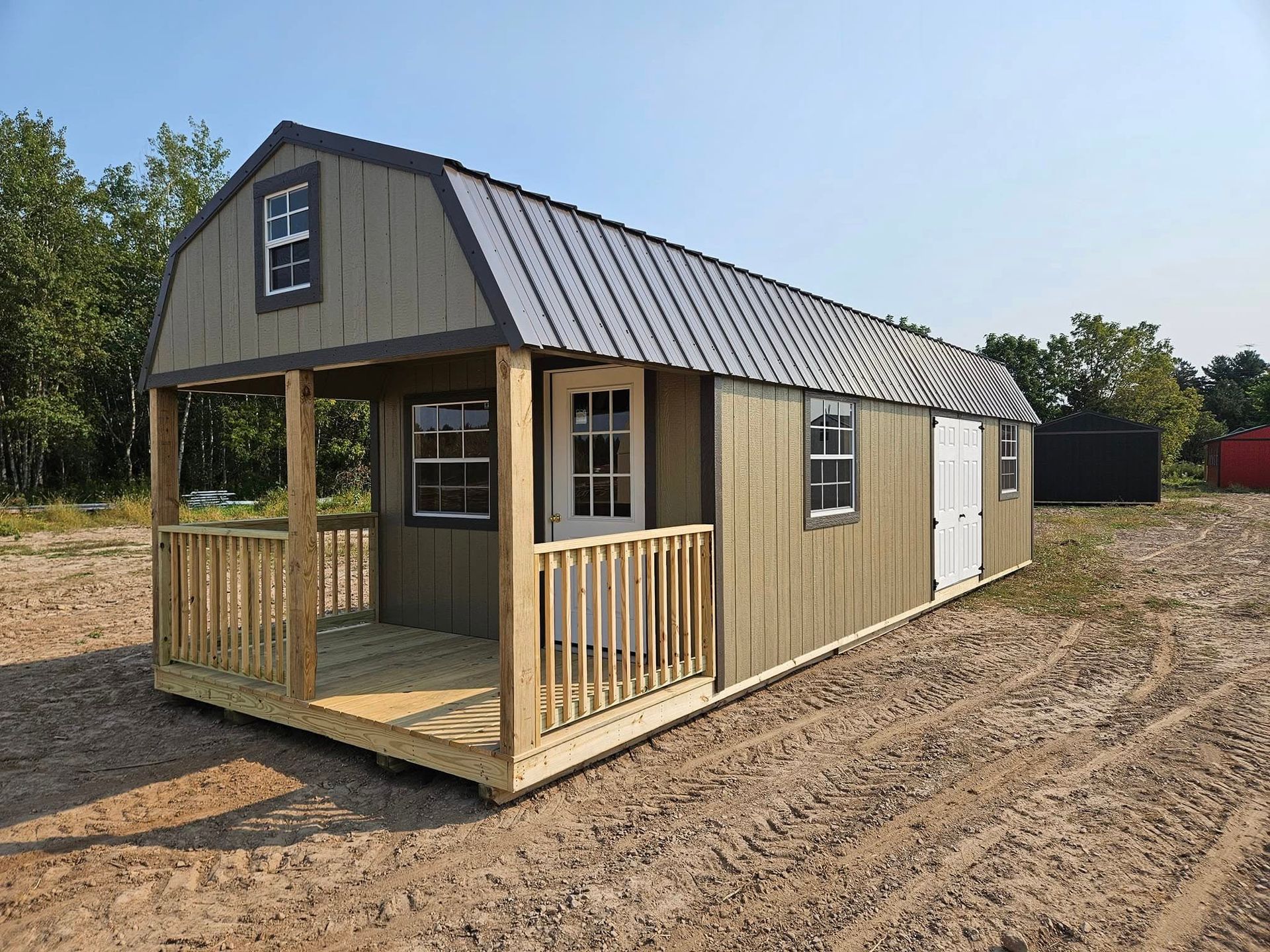 Small tan cabin-style tiny house with porch and dark roof on a dirt lot