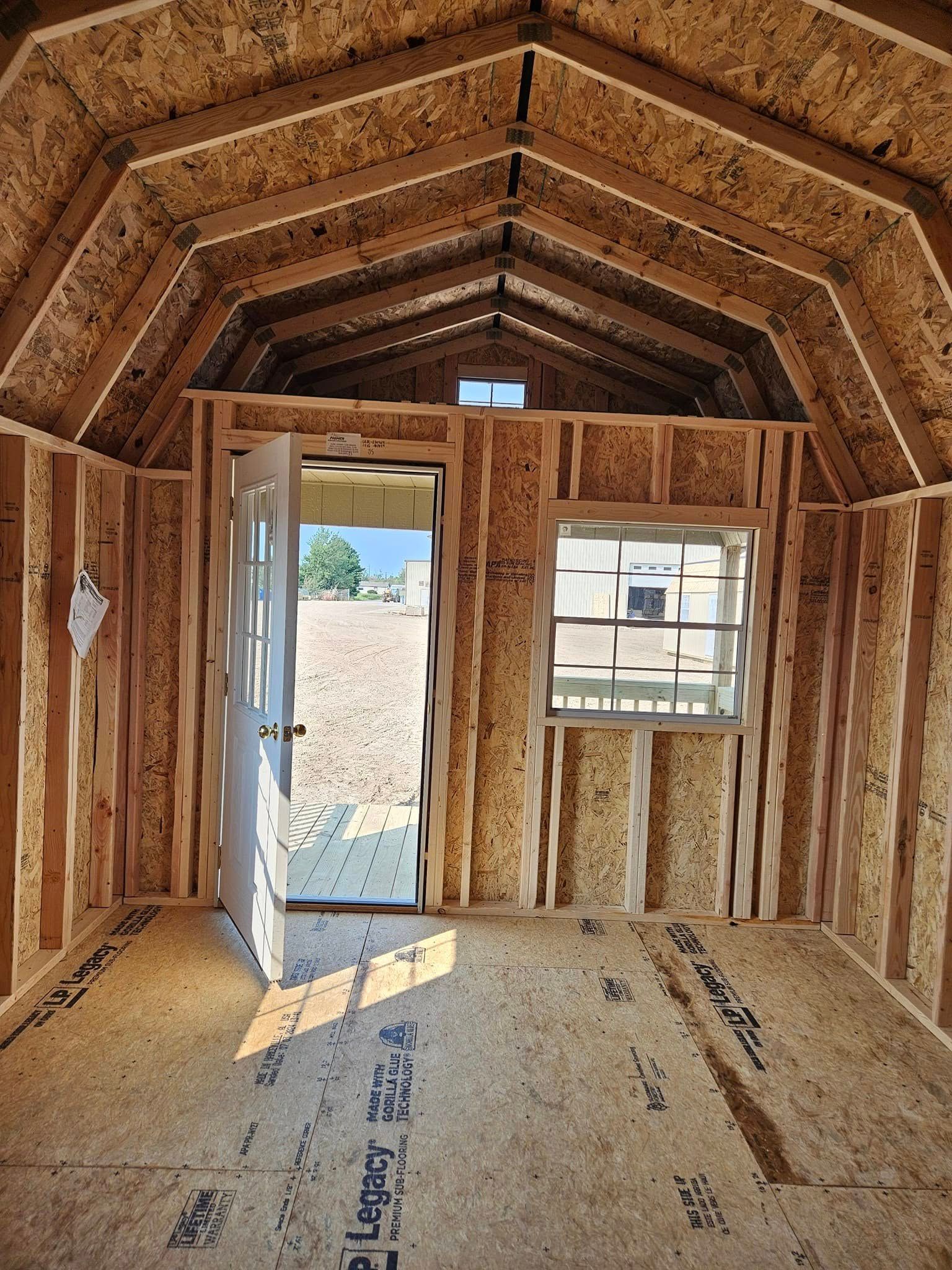 Interior of a small unfinished wooden shed with open doorway and two windows, sunlight streaming in.