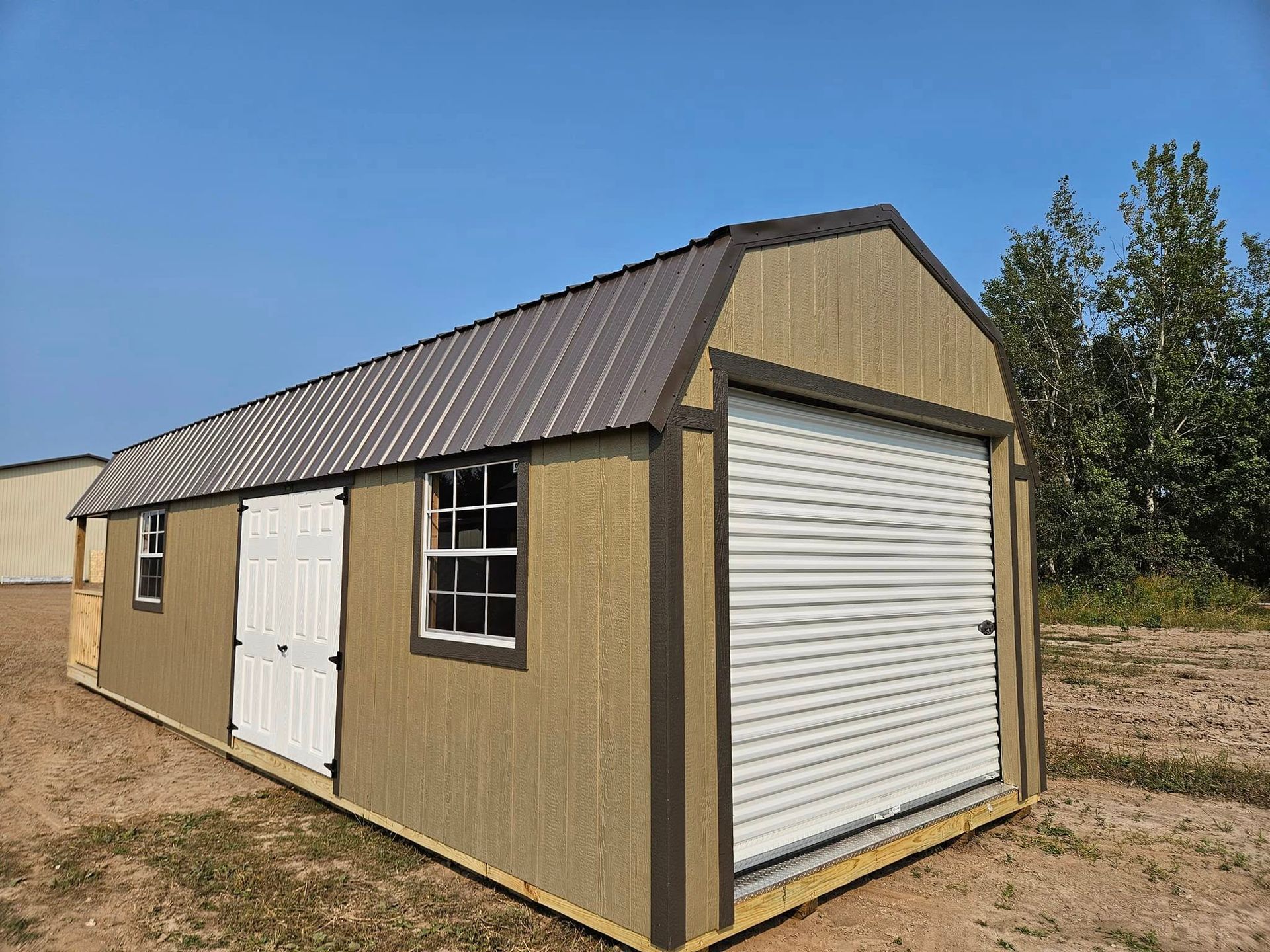 Tan metal storage shed with white roll-up door and side door in a grassy field under a blue sky