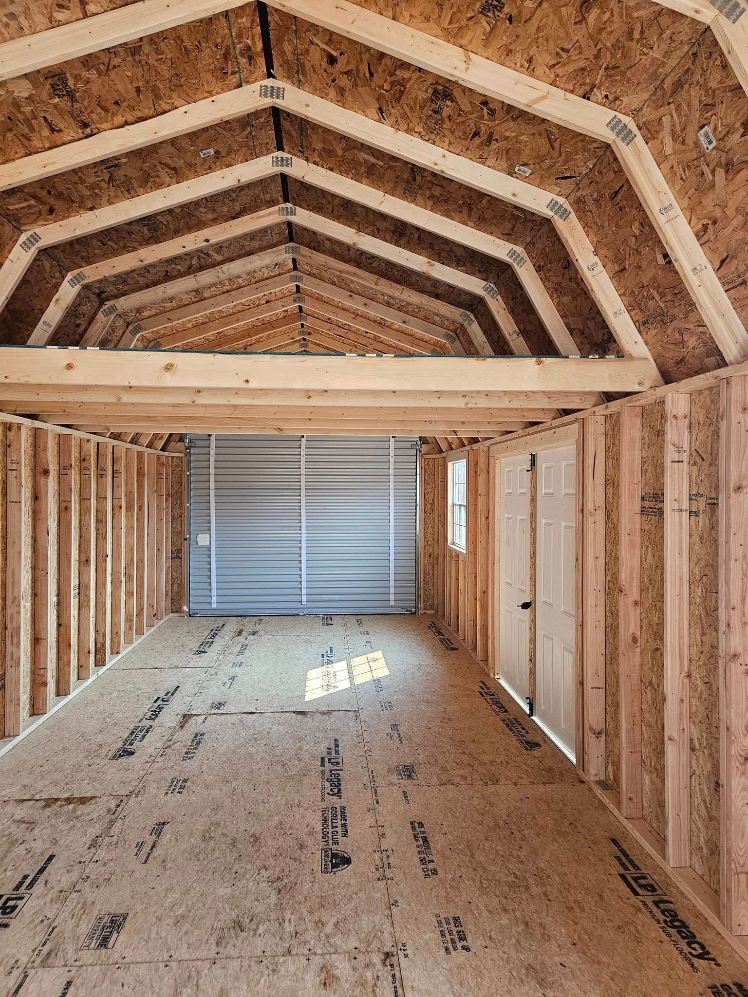 Interior of an unfinished wooden shed with exposed framing, plywood floor, and a closed roll-up door.