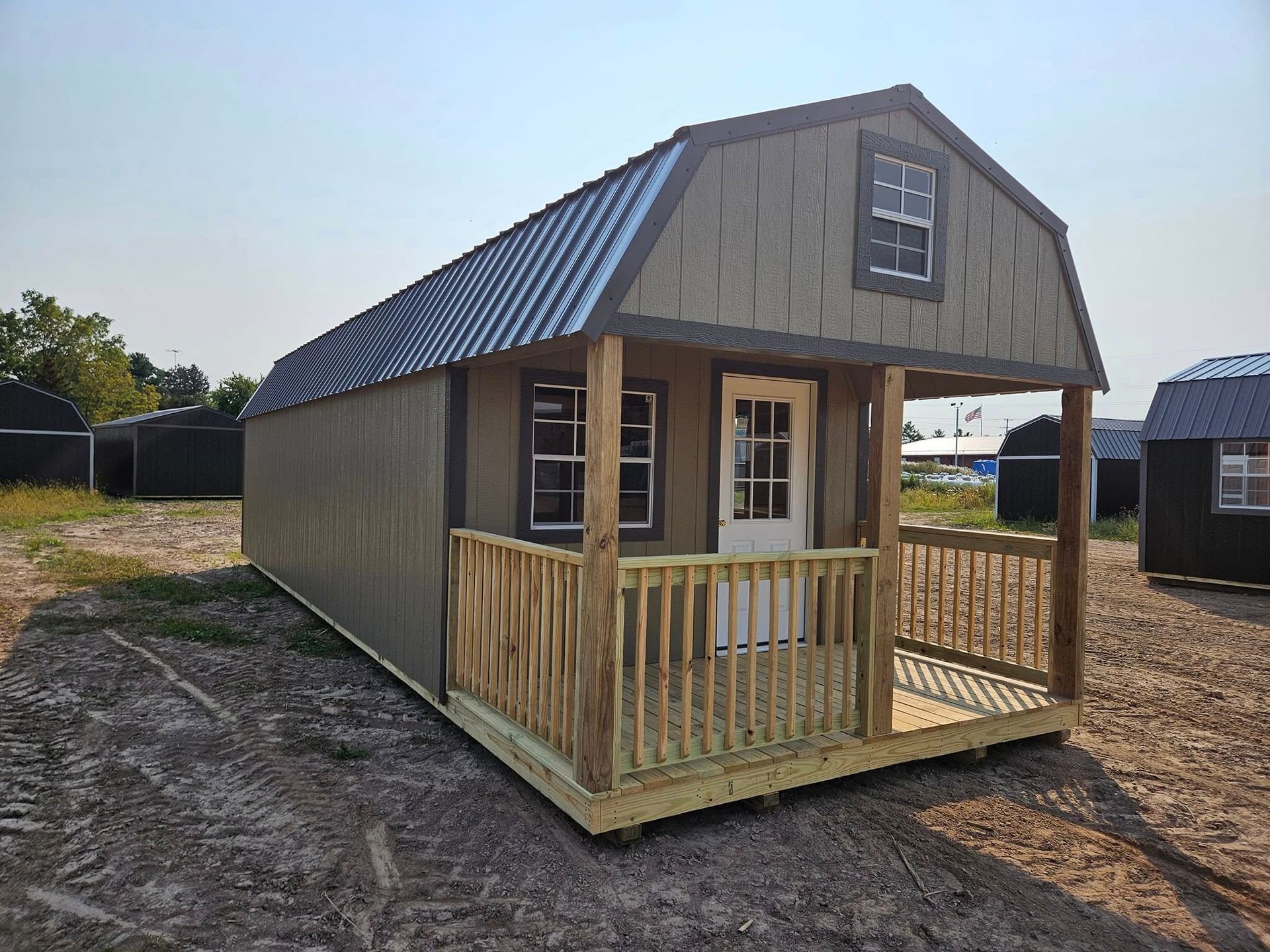Small tan shed-style cabin with a front porch, double doors, and a dark metal roof on a dirt lot.