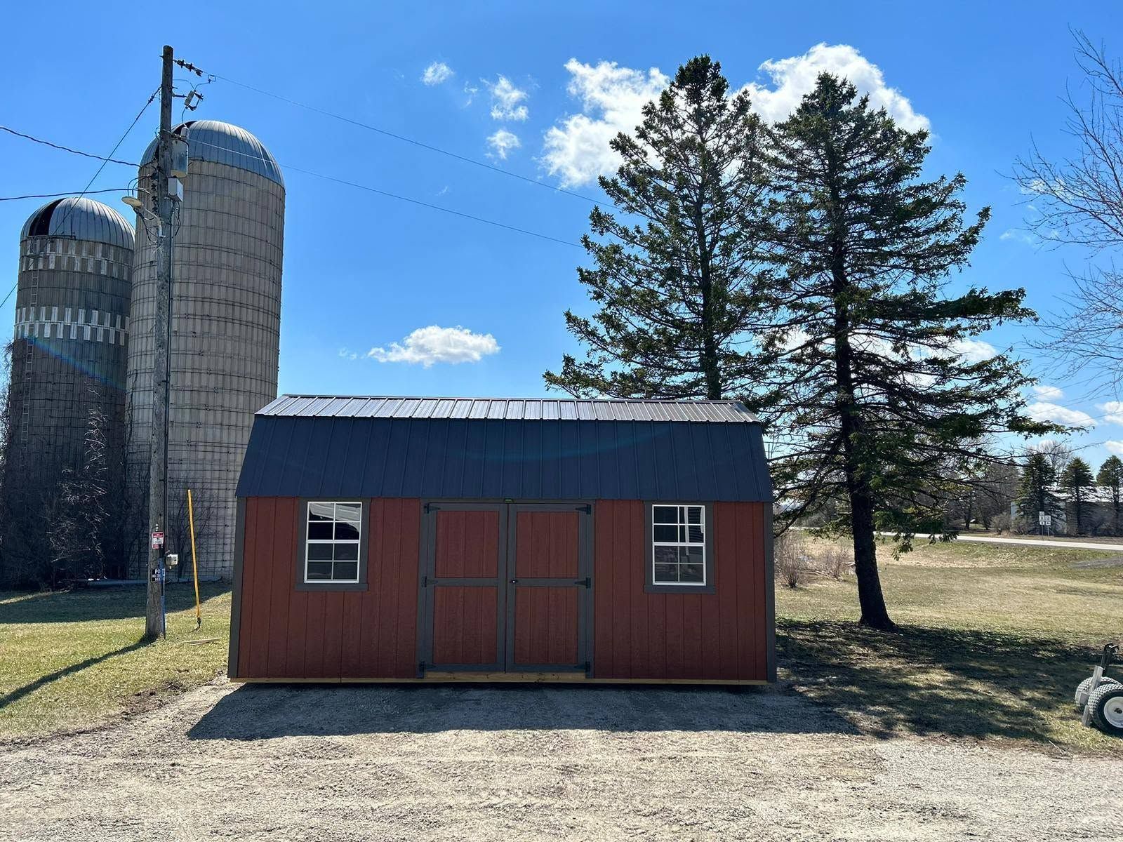 Red barn-style shed in a field beside a silo, under a blue sky with trees nearby.