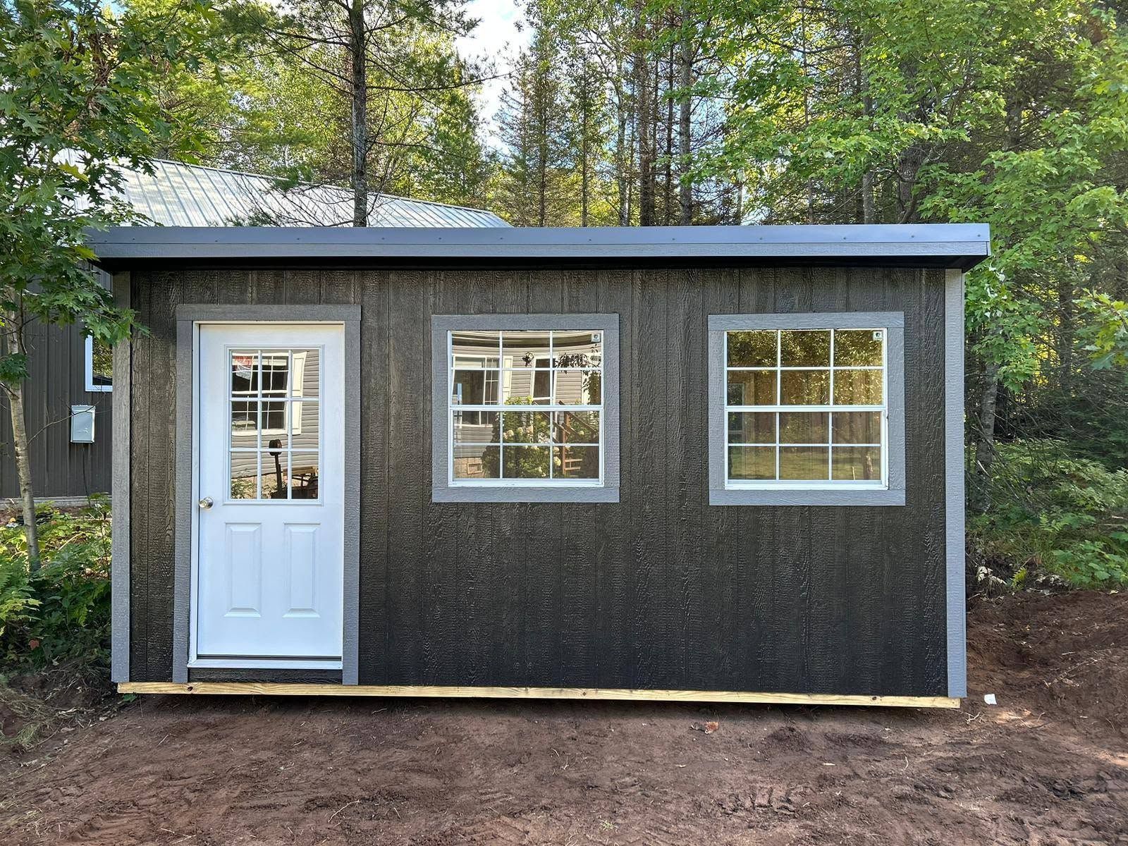 Small gray shed with white door and two windows, set in a wooded backyard