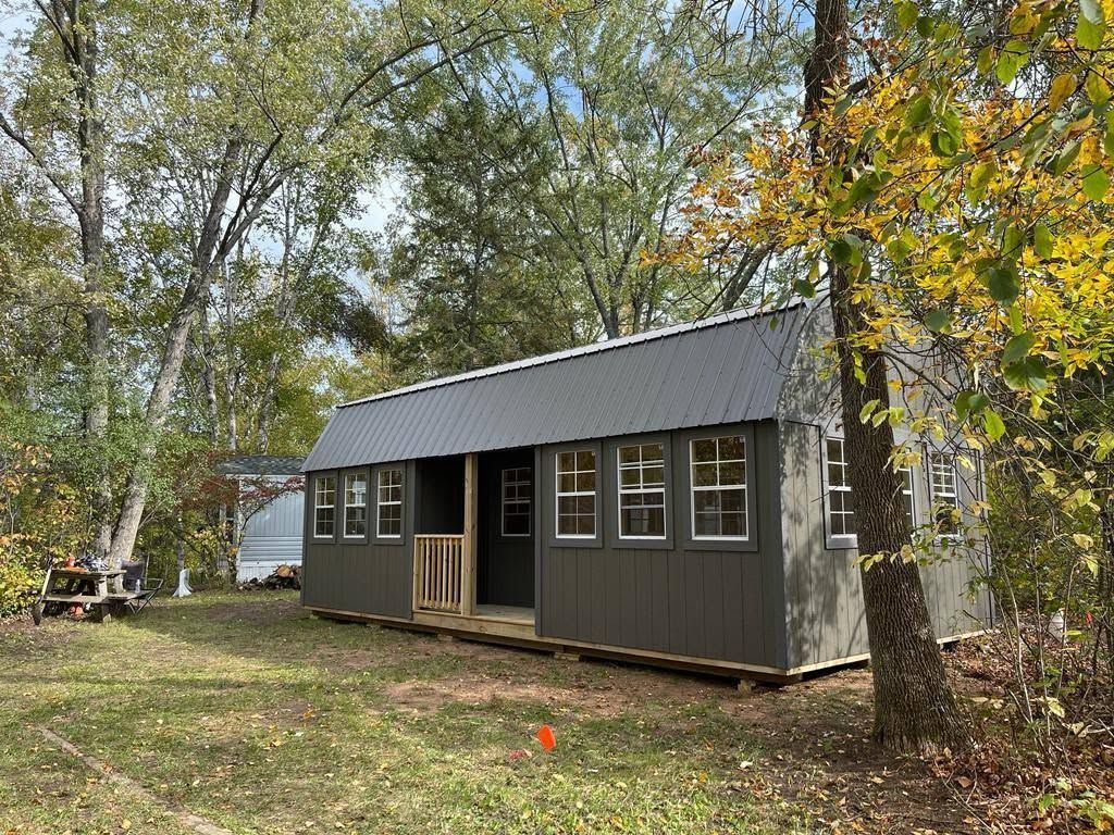 Small gray cabin with porch in a wooded yard, surrounded by trees and fallen leaves.