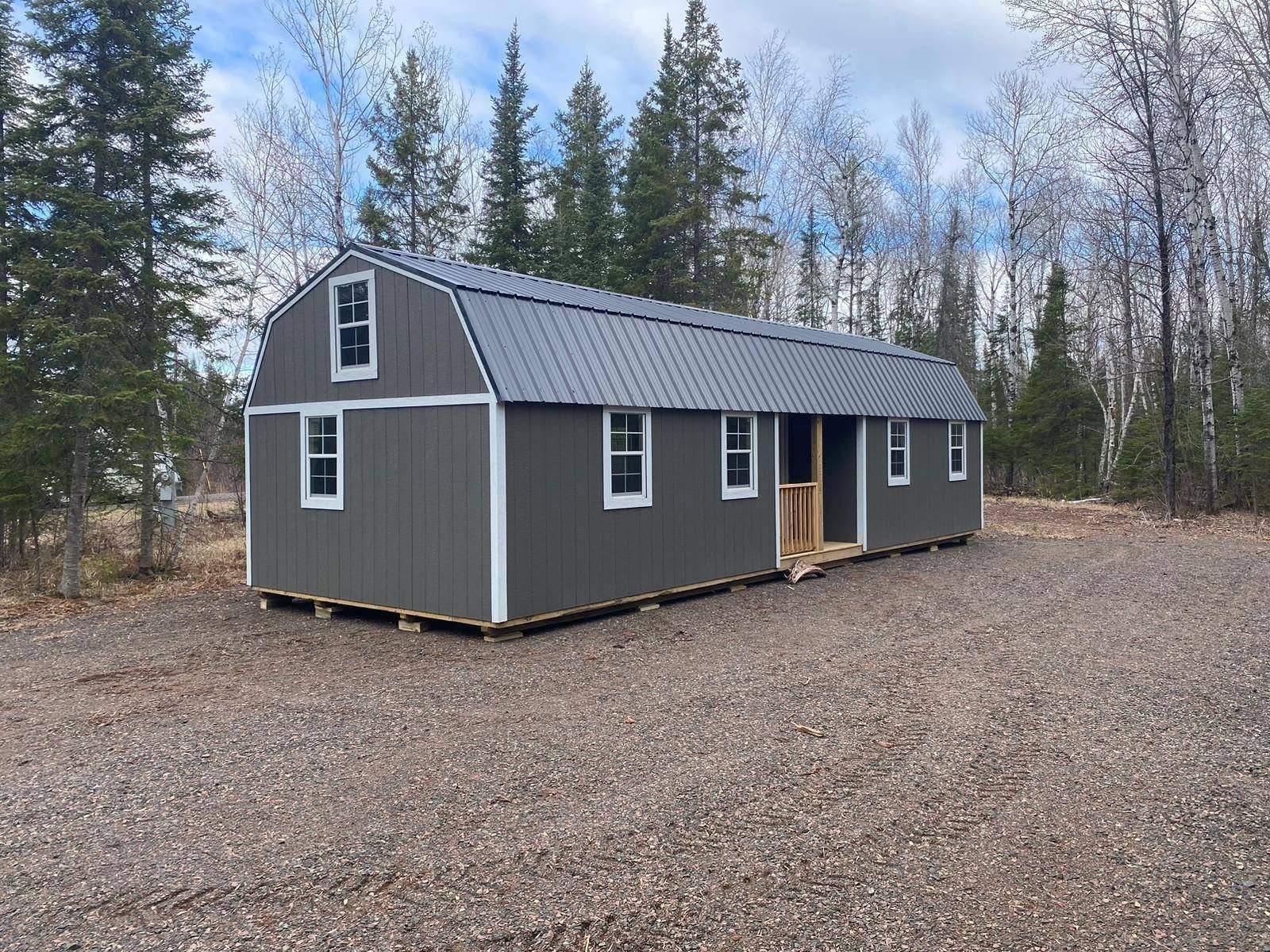 Small gray cabin with dark roof in a gravel clearing, surrounded by pine trees under a blue sky.