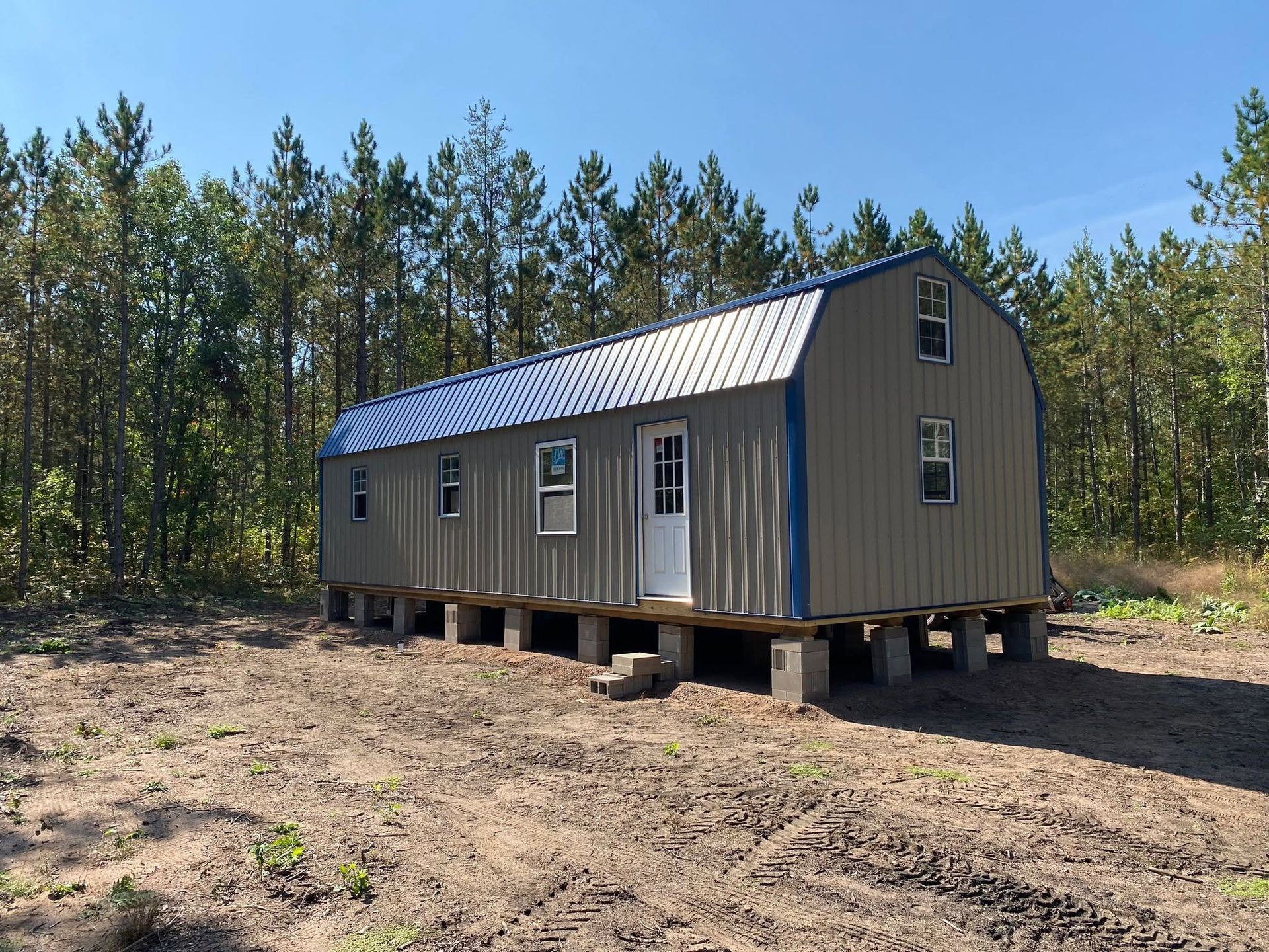 Portable cabin on blocks in a clearing, with tan siding, gray roof, and pine trees in the background